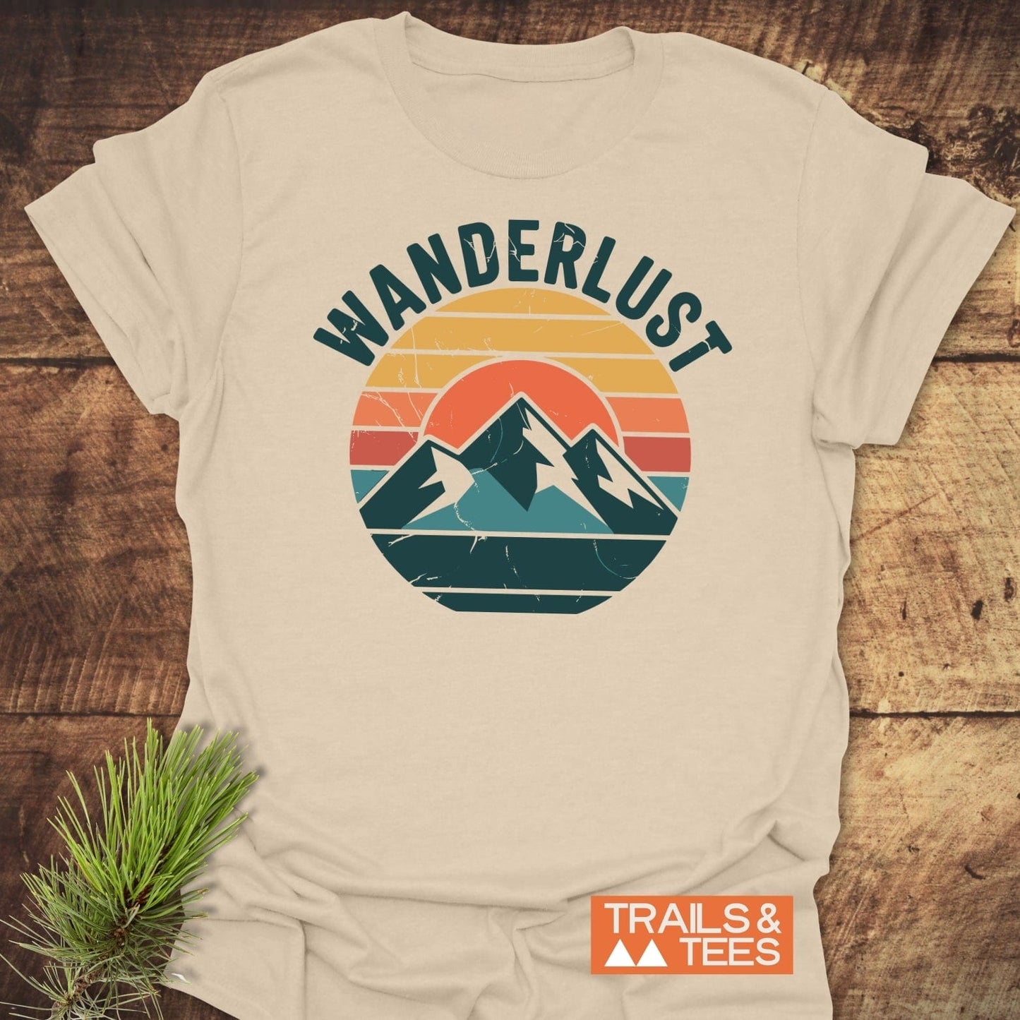 Wanderlust T-Shirt with retro mountain graphic and WANDERLUST text, made from soft, lightweight cotton, laid on wood with TRAILS & TEES logo and pine sprig.