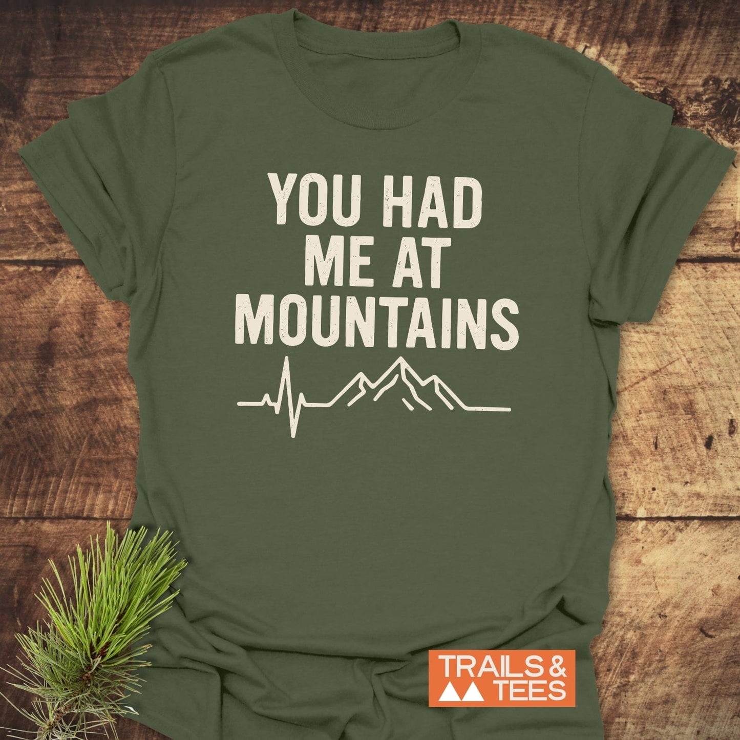 You Had Me At Mountains T-Shirt: army-green, off-white mountain graphic and text, orange “TRAILS & TEES” logo; laid on wooden surface with evergreen twig.