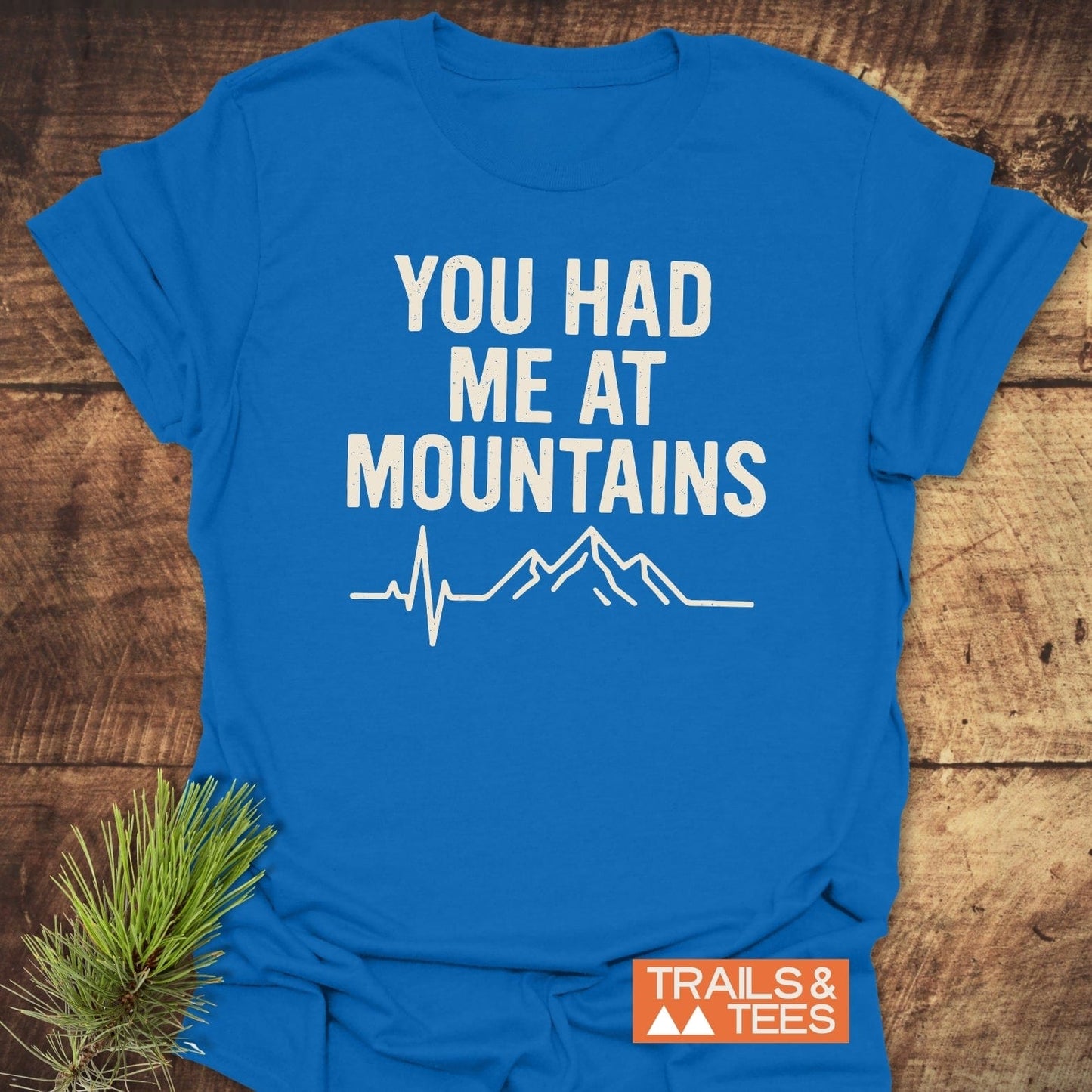 Flat-lay of the You Had Me At Mountains T-shirt with heartbeat and mountain design, featuring TRAILS & TEES logo, on a wooden surface with a pine sprig.
