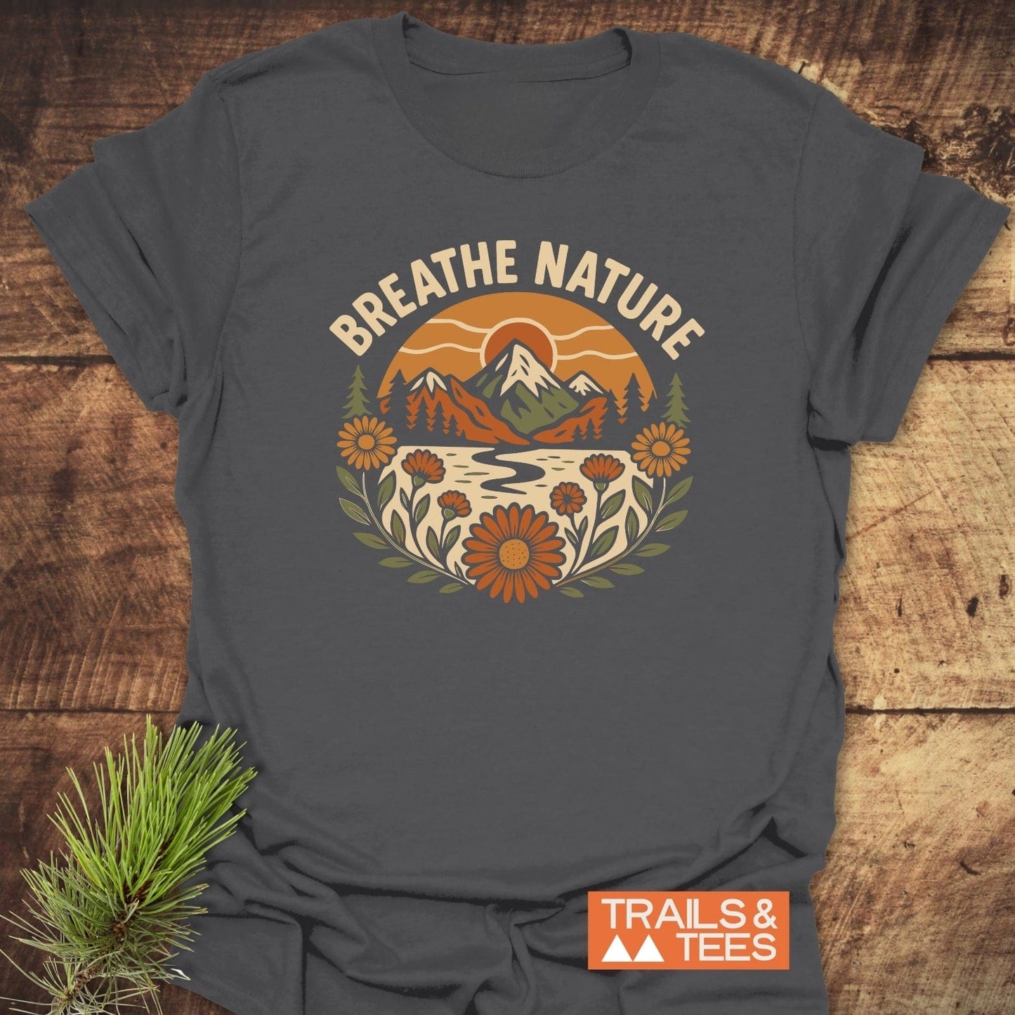 Breathe Nature T-Shirt on rustic wood, features retro nature graphic with mountains and wildflowers, showcasing a slogan. Pine sprig accentuates outdoor theme.