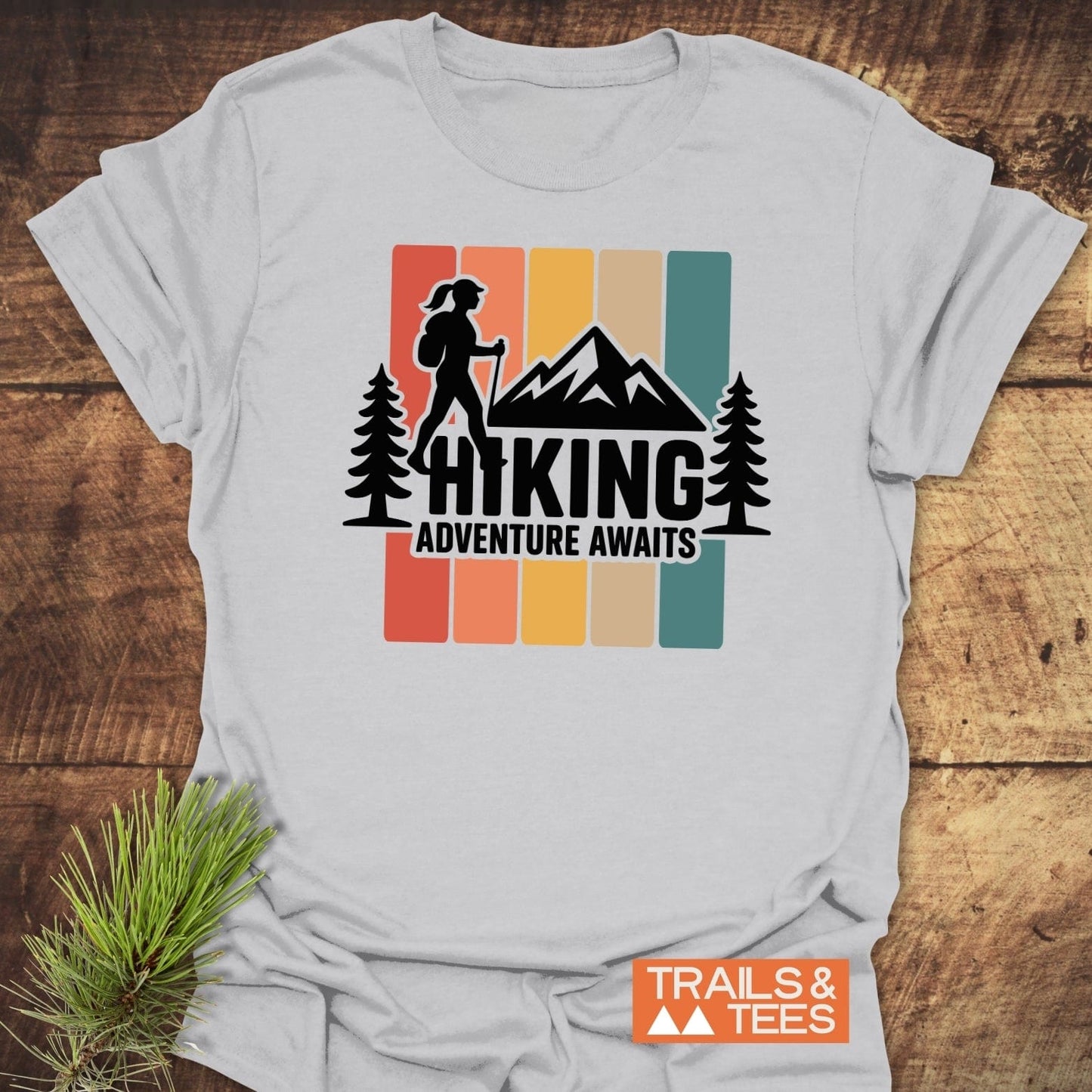 Hiking Girl T-Shirt with retro color blocks, hiker silhouette, and HIKING ADVENTURE AWAITS text, laid on a wooden surface with TRAILS & TEES logo.