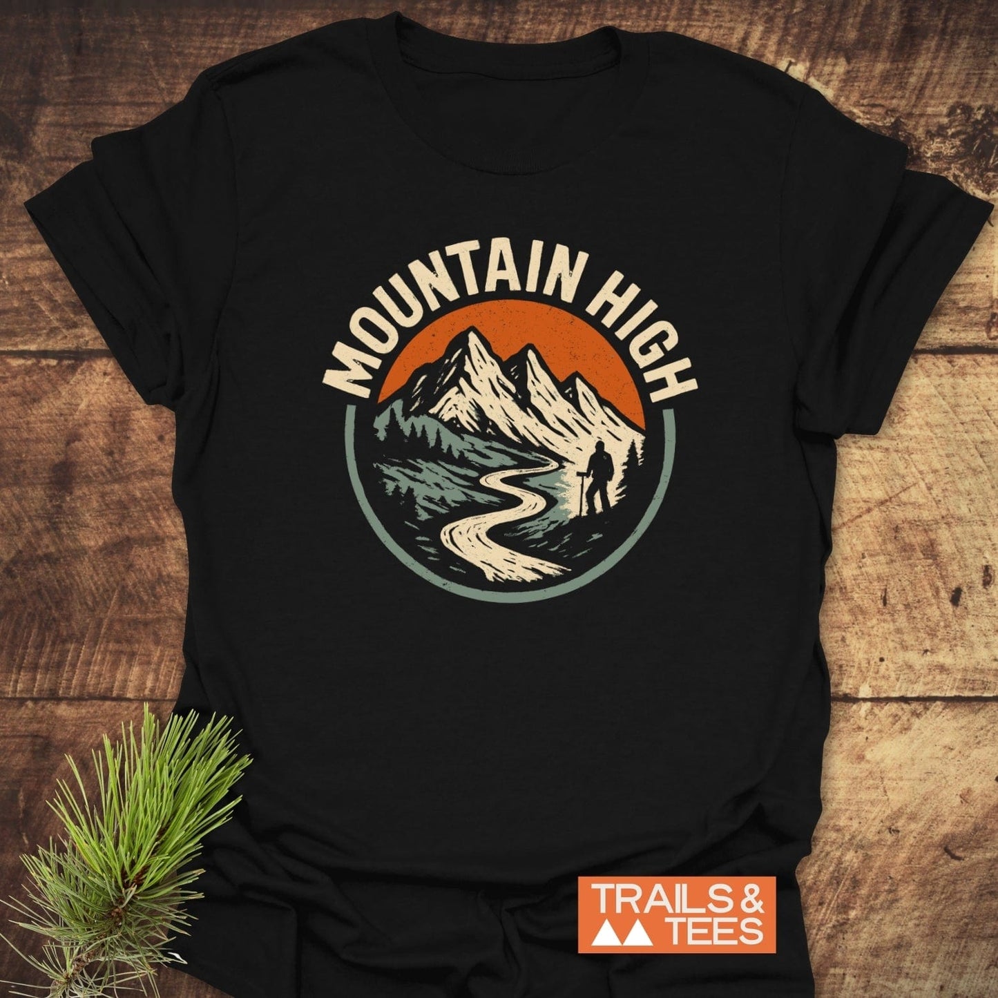 Mountain High T-Shirt featuring a vintage mountain graphic with a hiker, crafted from soft ring-spun cotton for comfort and durability, next to TRAILS & TEES logo.
