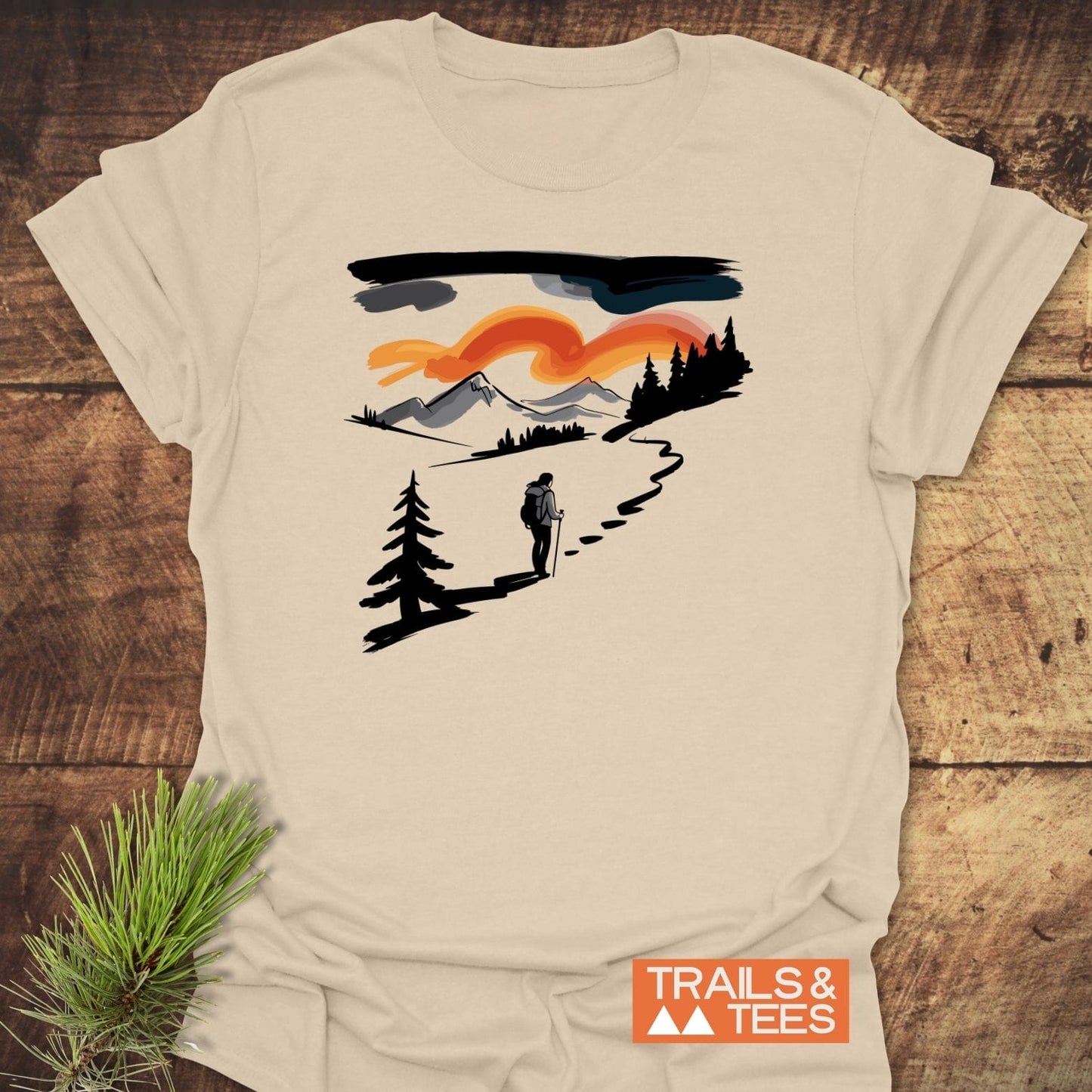 Minimalist Ink Hiking T-Shirt featuring a hiker on a trail with mountain and sunset backdrop, showcasing outdoor adventure spirit, laid flat beside a pine branch.