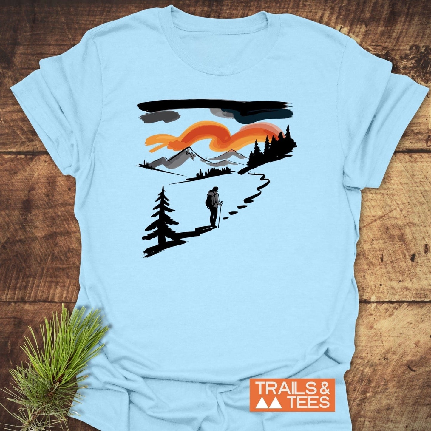 Minimalist Ink Hiking T-Shirt on rustic wood, featuring a hiker graphic with mountains and trees. Made from soft cotton, ideal for outdoor enthusiasts.