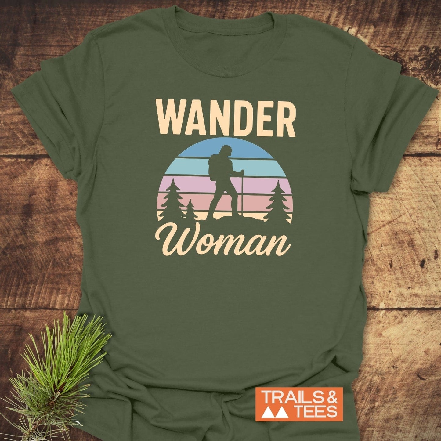 Wander Woman Retro T-Shirt with sunset graphic and hiker print, on rustic wood, alongside pine sprig; features TRAILS & TEES logo, crafted for outdoor adventures.