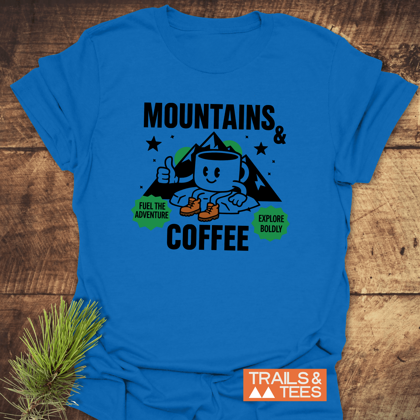 Mountains & Coffee Retro T-Shirt featuring a hiking coffee mug graphic, mountain peaks, and motivational badges, laid on wood with TRAILS & TEES branding.