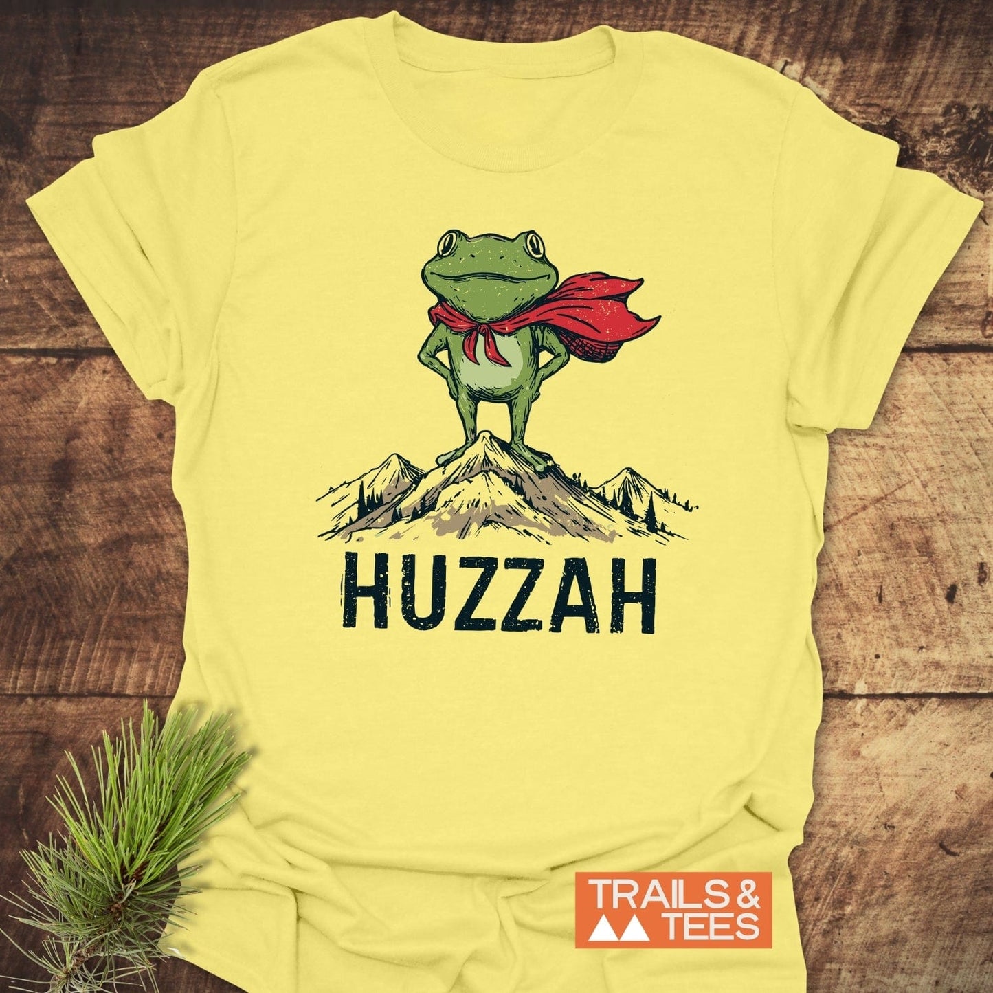 Trail Hero Funny T-Shirt featuring a cartoon frog in a red cape on a mountain with HUZZAH text, showcasing durable, comfortable, and eco-friendly design.