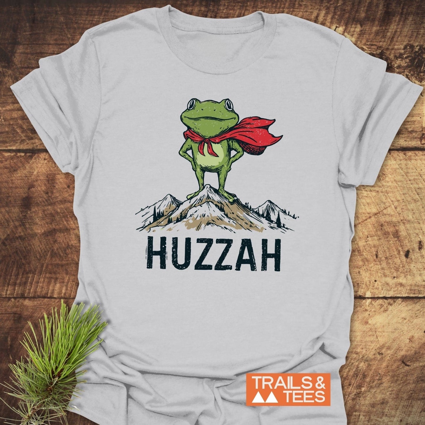 Trail Hero Funny T-Shirt featuring a cartoon green frog in a red cape atop a mountain, with “HUZZAH” text, showcasing adventure-ready design and comfort.