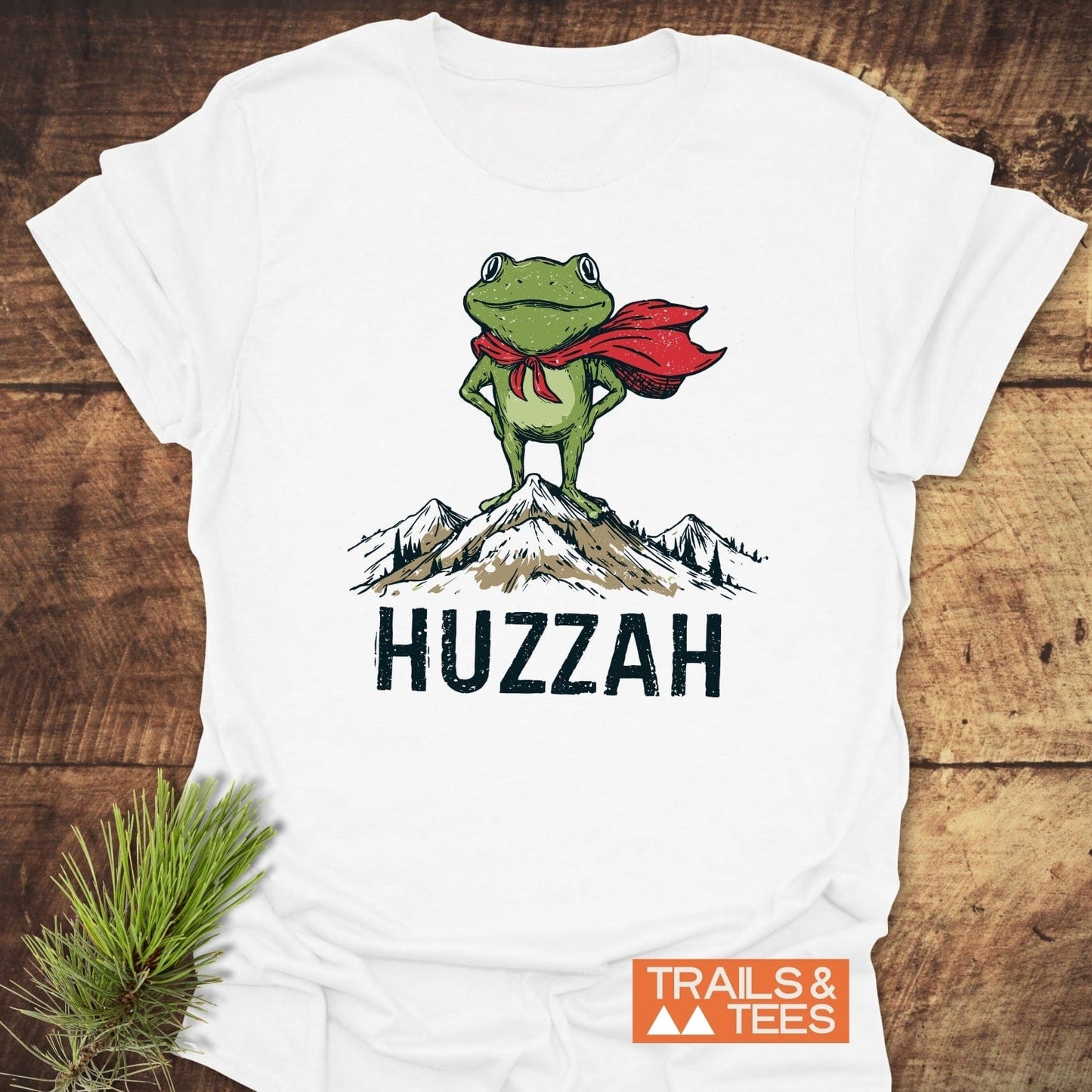 Trail Hero Funny T-Shirt with a frog in a cape on a mountain, HUZZAH text, and TRAILS & TEES logo, laid on wood with pine sprig.