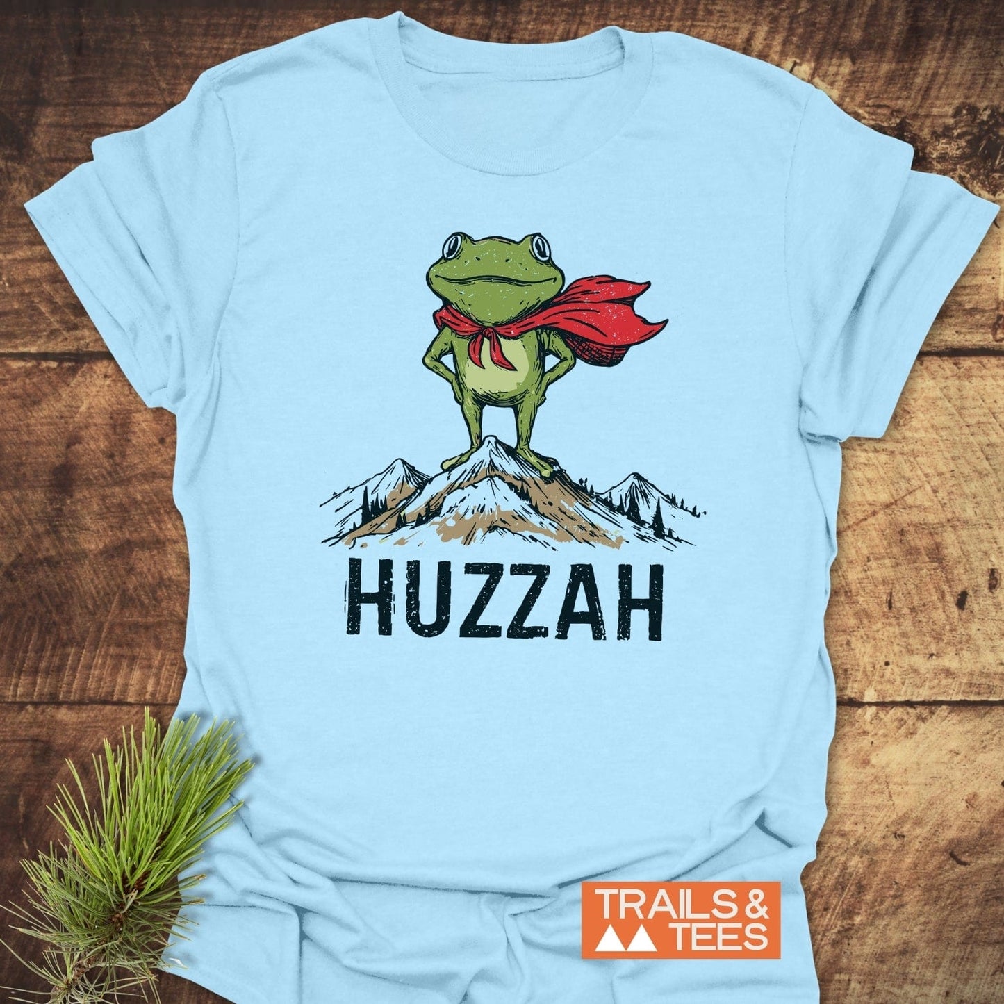 Trail Hero Funny T-Shirt featuring a cartoon frog in a cape on a mountain, with HUZZAH text and TRAILS & TEES logo, laid on wood.