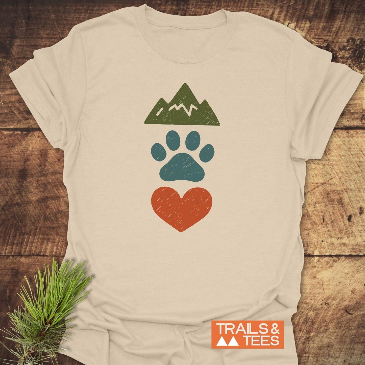 Mountains & Dogs Love T-Shirt on wood, featuring mountain, paw print, and heart graphics. Made of soft, lightweight cotton for outdoor adventures. Brand: TRAILS & TEES.