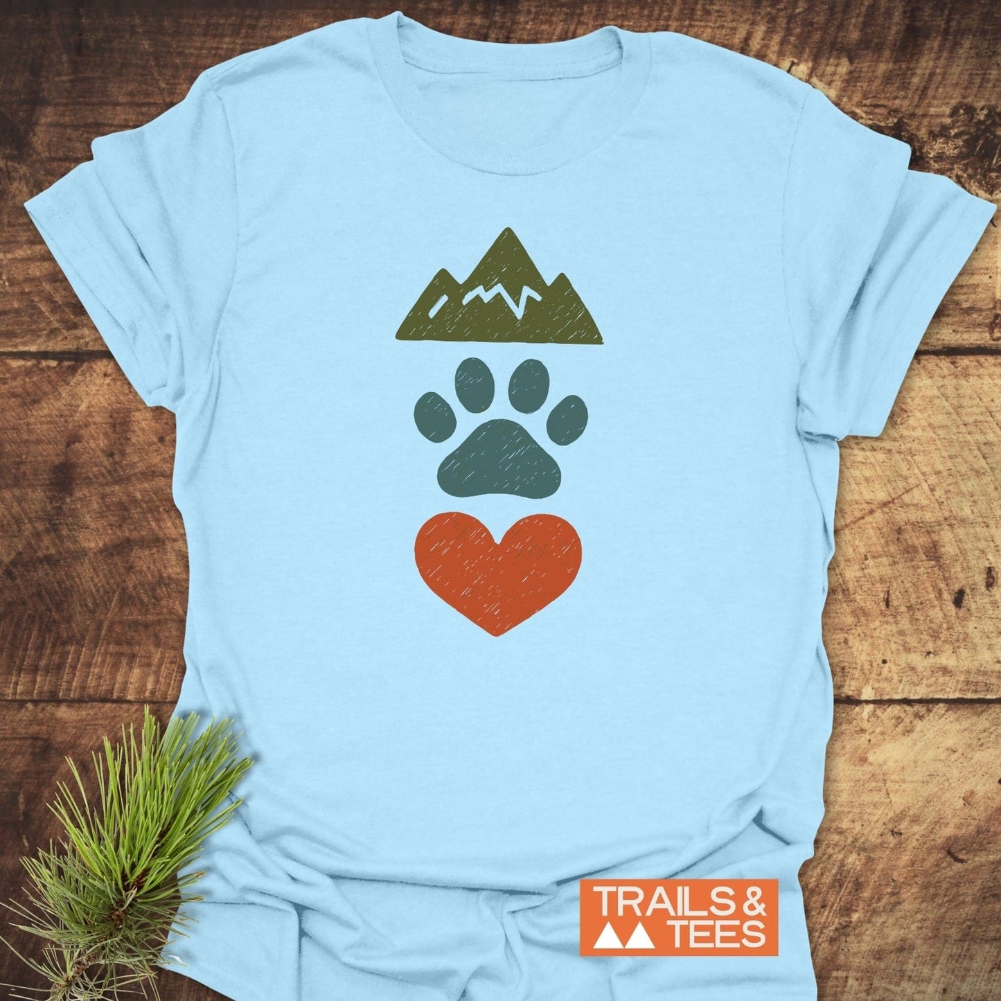 Mountains & Dogs Love T-Shirt featuring mountain, paw, and heart icons, laid on wood with pine sprig. Includes Trails & Tees logo.
