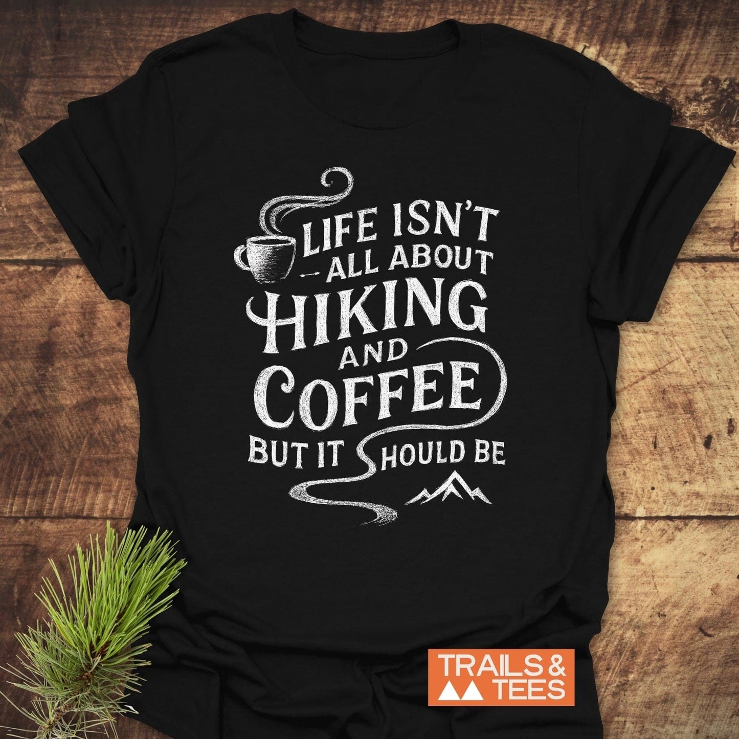 Black Hiking and Coffee T-Shirt featuring a white chalk-style design with a coffee cup, hiking trail, and mountains, laid flat on wood.