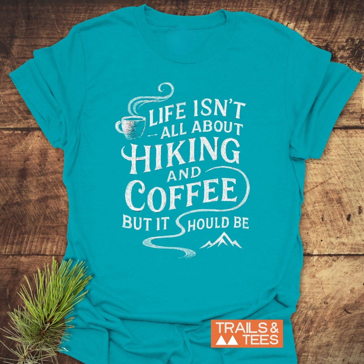 Hiking and Coffee T-Shirt with a slogan featuring a steaming coffee cup and mountains, laid on wood, includes TRAILS & TEES logo in the corner.