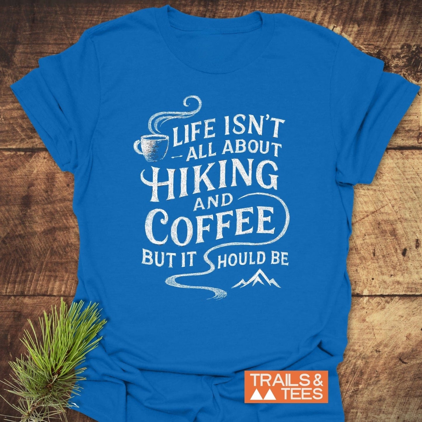Hiking and Coffee T-Shirt featuring a coffee cup, slogan, and mountain graphic, laid on wood with pine sprigs; soft, durable cotton for outdoor enthusiasts.
