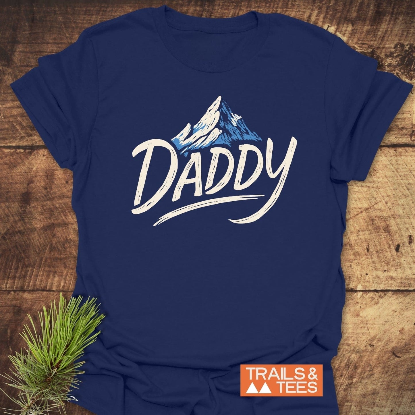 Mountain Daddy T-Shirt with snow-topped mountain illustration and “Daddy” text, laid on rustic wood with a pine sprig, emphasizing its outdoor theme and comfort.