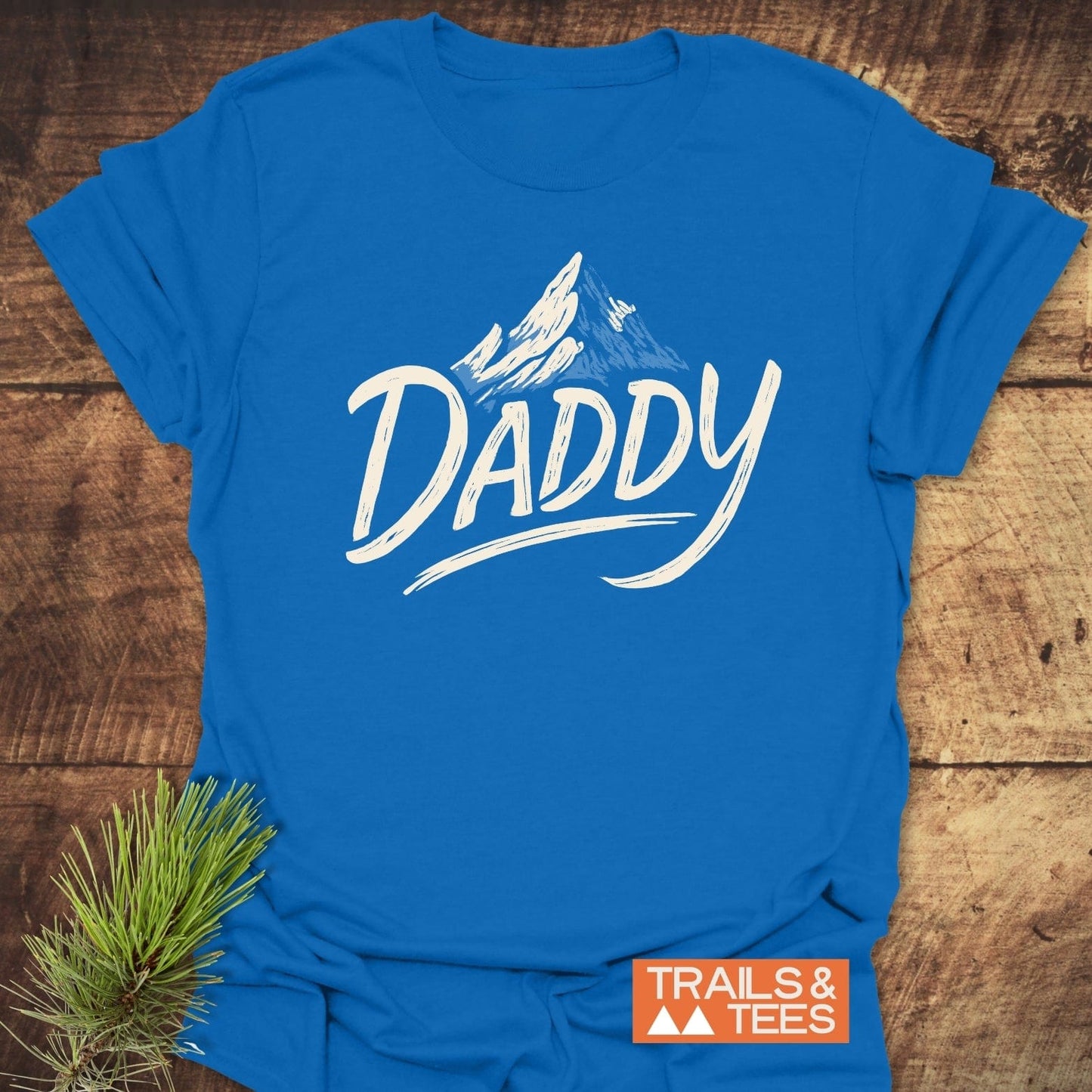 Mountain Daddy T-Shirt featuring Daddy script with mountain sketch. Made of soft, ring-spun cotton, ensuring comfort and durability. Includes Trails & Tees logo.