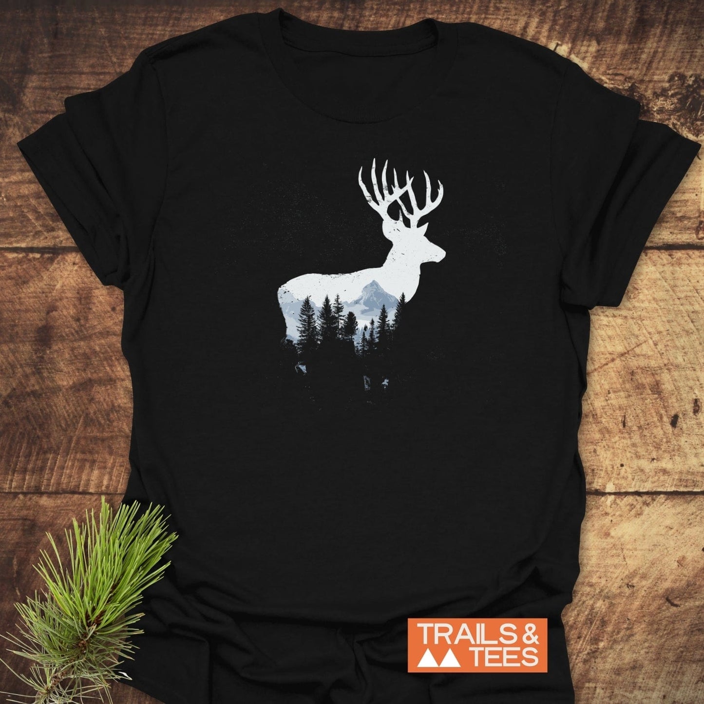 Deer Silhouette T-Shirt featuring a stag with a mountain scene print on black cotton. Laid on wood with logo and pine sprig visible.