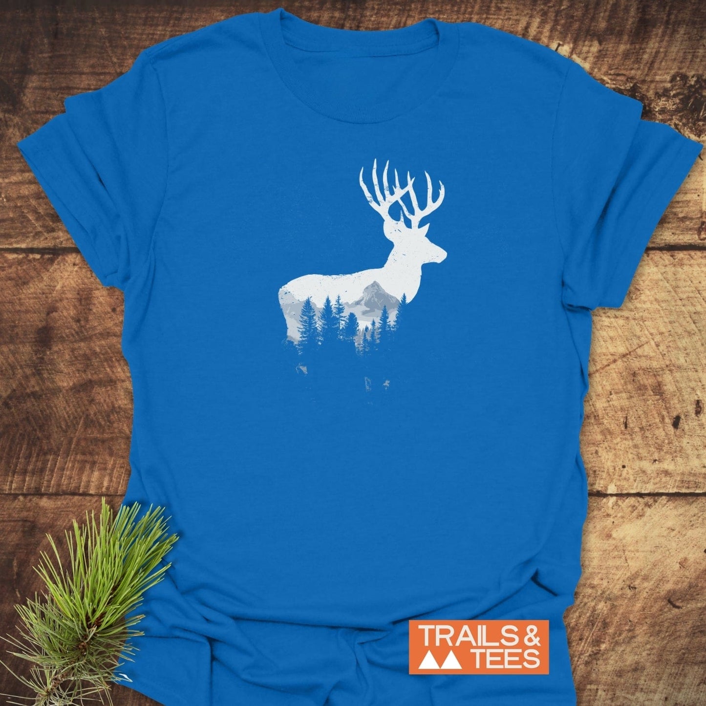Deer Silhouette T-Shirt with a stag design featuring a mountain landscape, placed on wood. Includes “TRAILS & TEES” label and pine needle accent.