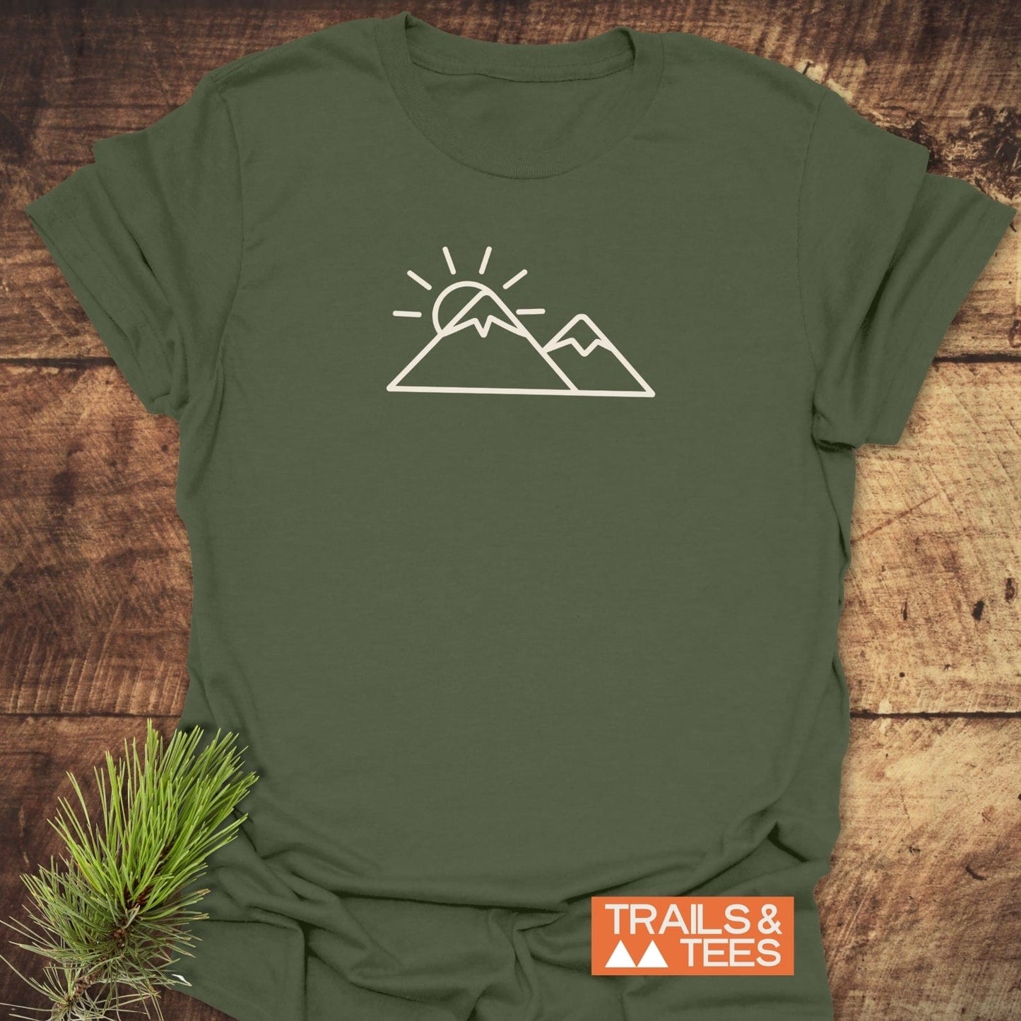 Minimalist Mountains And Sun T-Shirt featuring a white mountain and sun design, TRAILS & TEES logo, laid flat on wood with pine sprig.