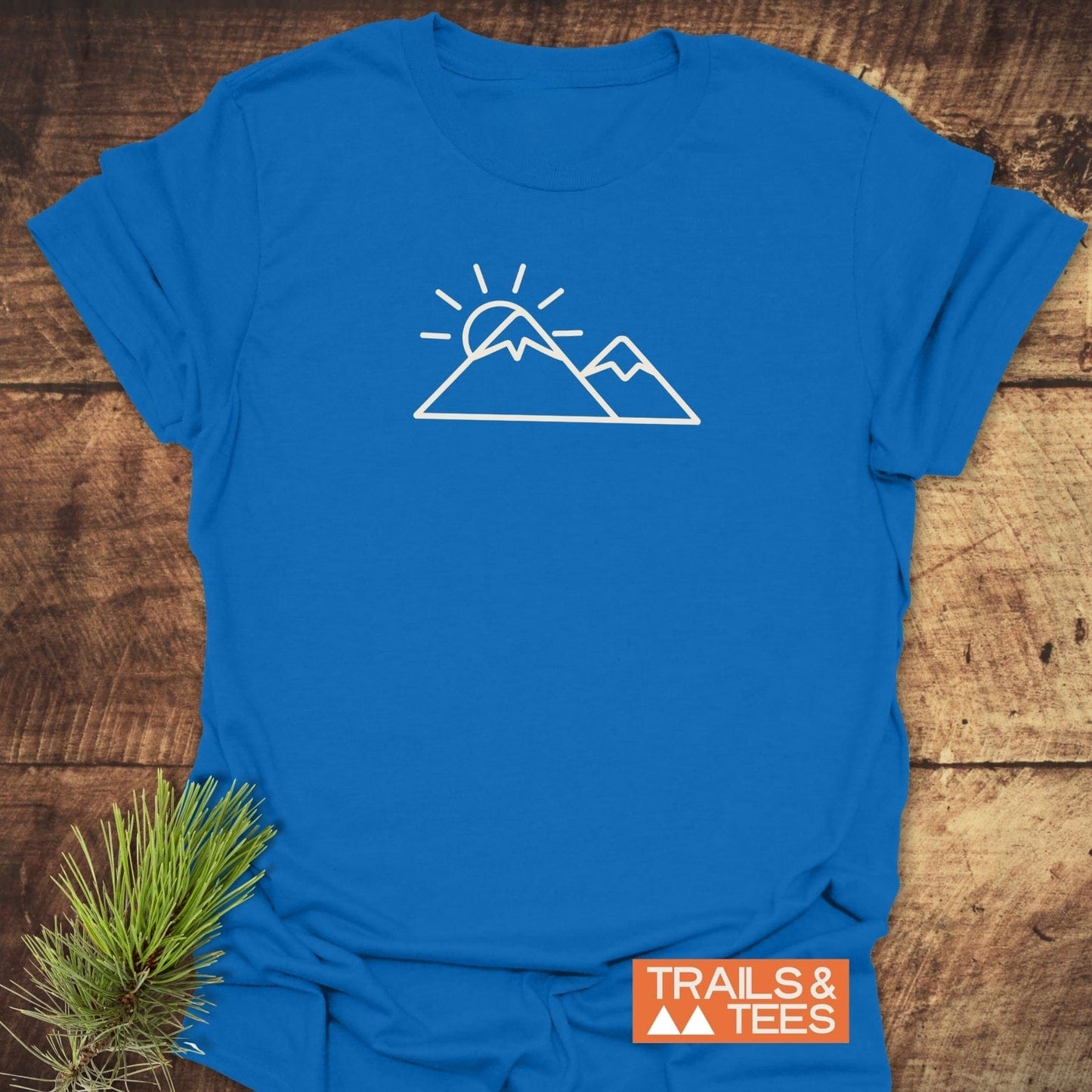Minimalist Mountains And Sun T-Shirt featuring a white mountain and sun graphic on blue cotton, displayed with a pine sprig and “TRAILS & TEES” logo.