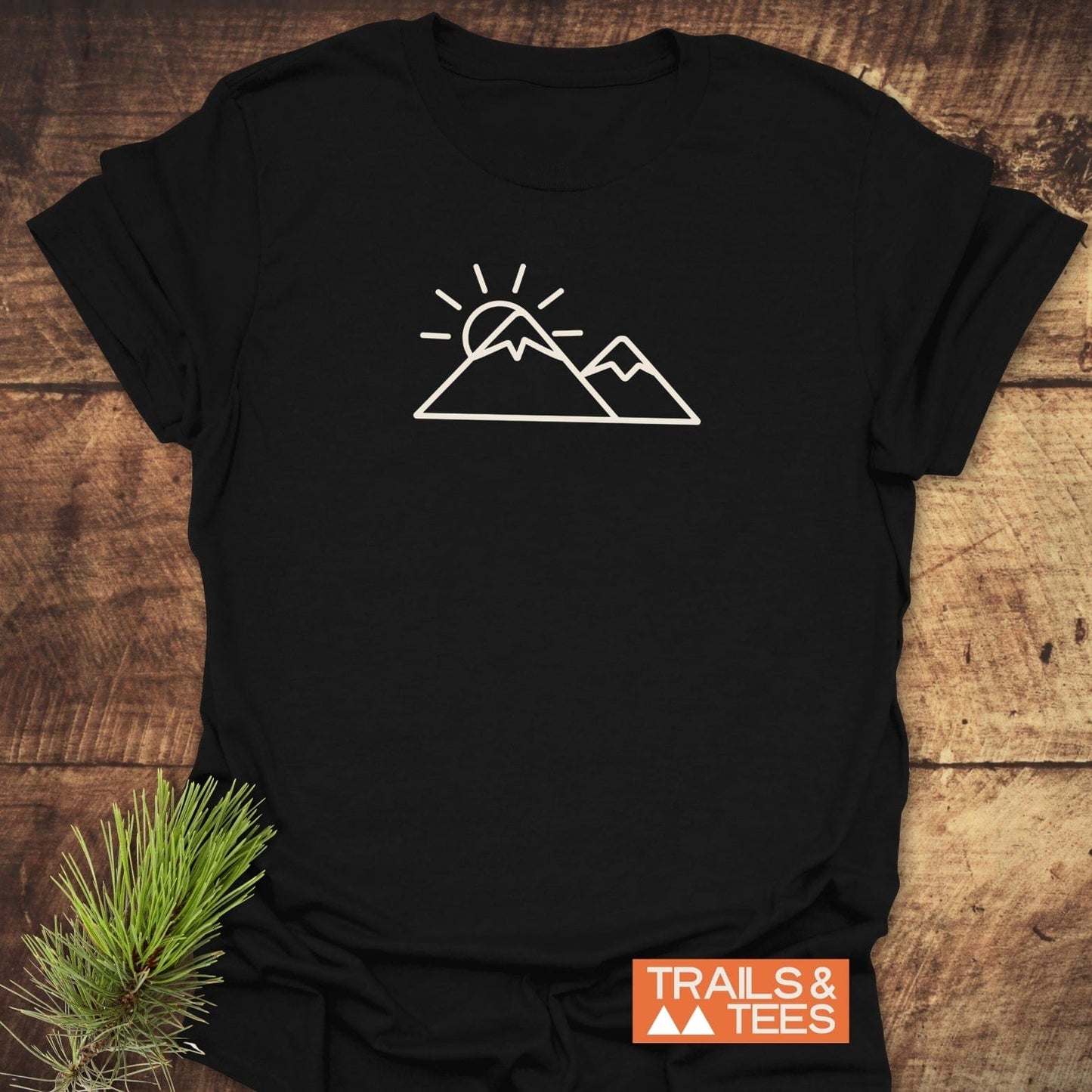 Minimalist Mountains And Sun T-Shirt on rustic wood, featuring a white line-drawing of mountains and sun. Includes orange TRAILS & TEES label.