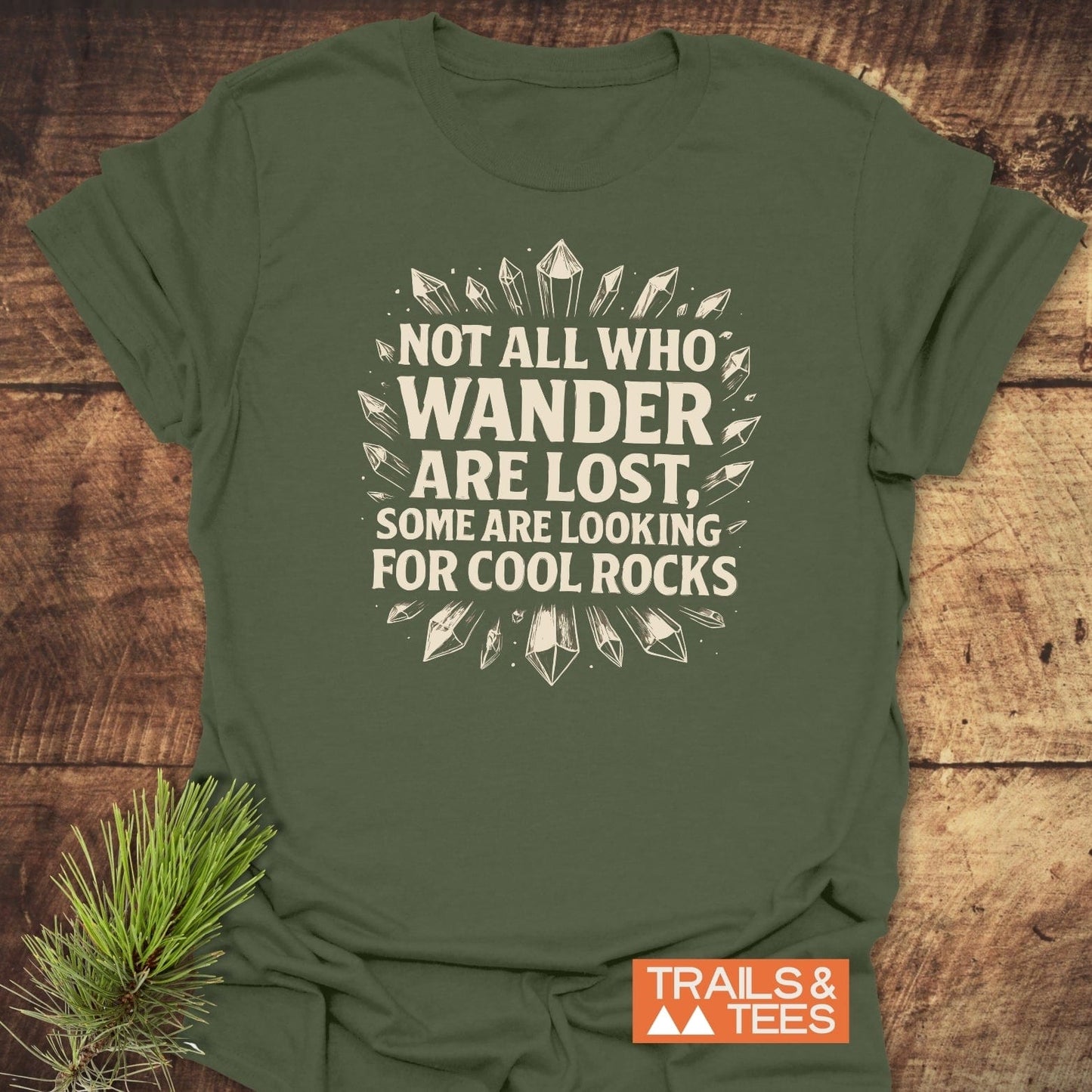 Olive-green 764 Not All... Rock Lover T-Shirt with rock illustrations, slogan in off-white, on wooden surface, beside a pine sprig, Trails & Tees logo visible.