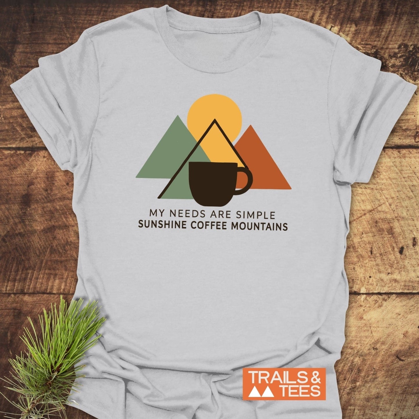 The Simple Joys T-Shirt features a mountain graphic with a sun and coffee mug, slogan, and Trails & Tees branding on light-gray fabric.