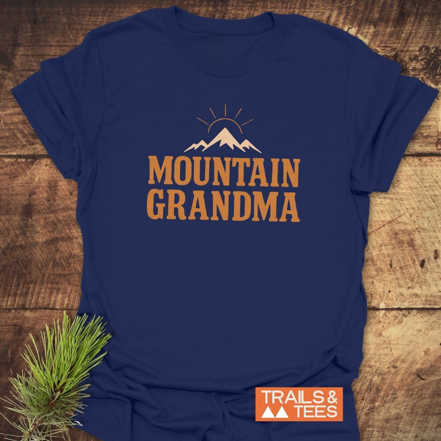 Mountain Grandma T-Shirt featuring a mountain graphic with rising sun on navy-blue, laid on wood. Includes “TRAILS & TEES” logo and pine sprig.