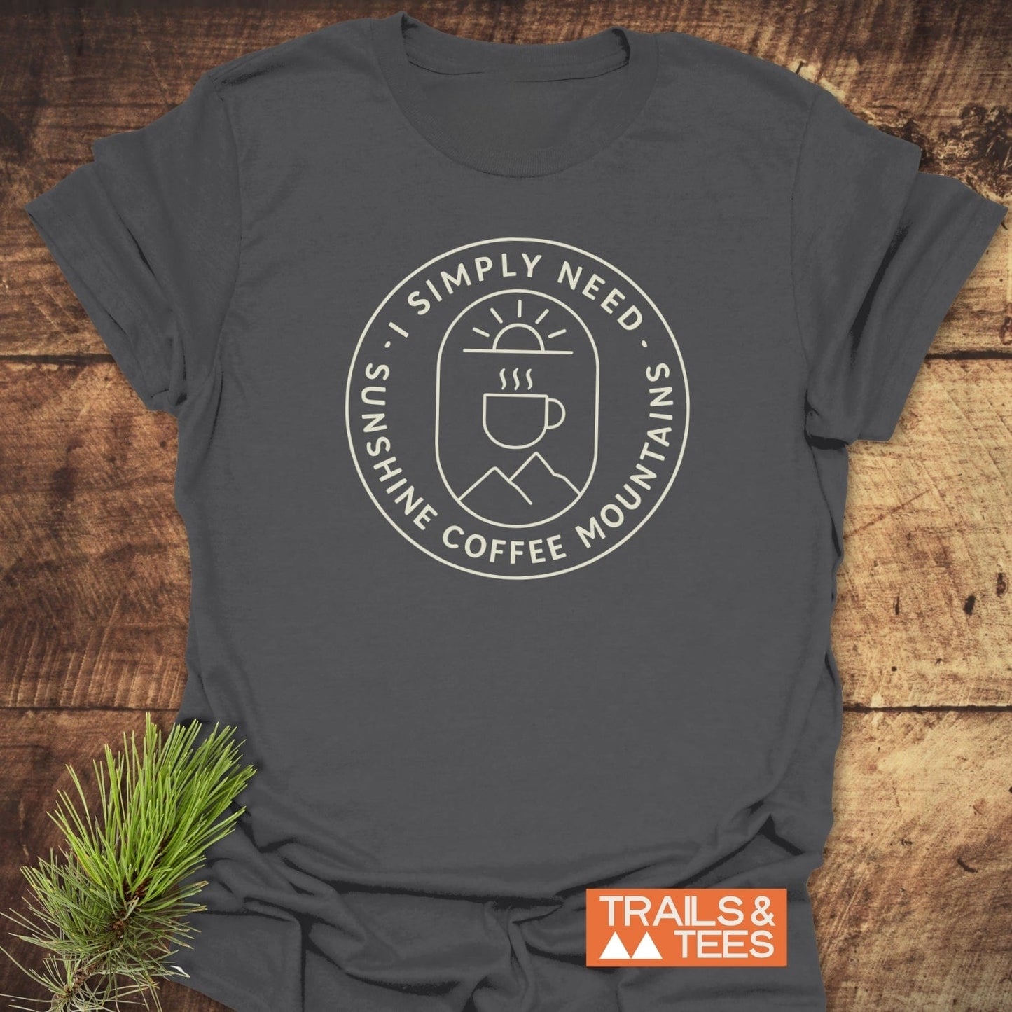 Minimalist Sunshine, Coffee & Mountains T-Shirt with circular graphic of sun, coffee, and mountains; slogan encircles image. Trails & Tees logo and evergreen sprig beside.