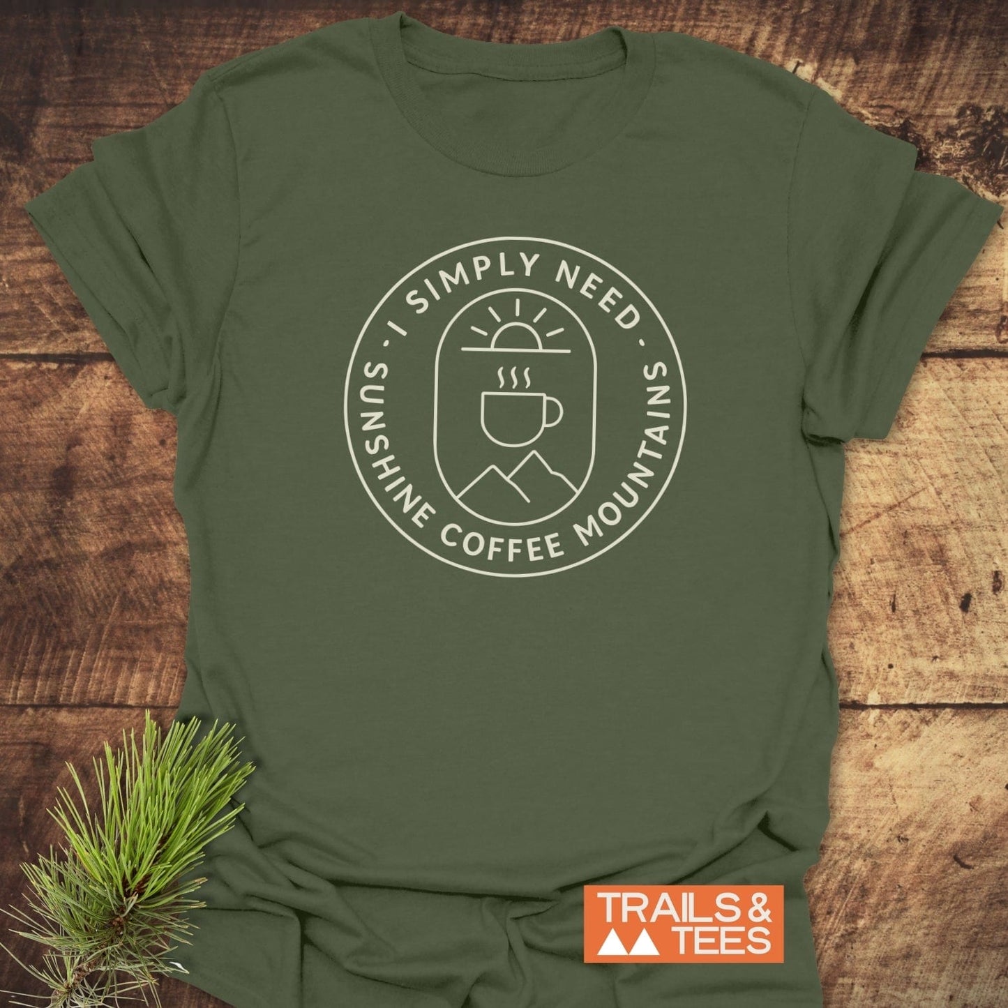 Minimalist Sunshine, Coffee & Mountains T-Shirt featuring a circular sunrise and mountain graphic on forest-green cotton, laid on rustic wood with a pine sprig beside it.