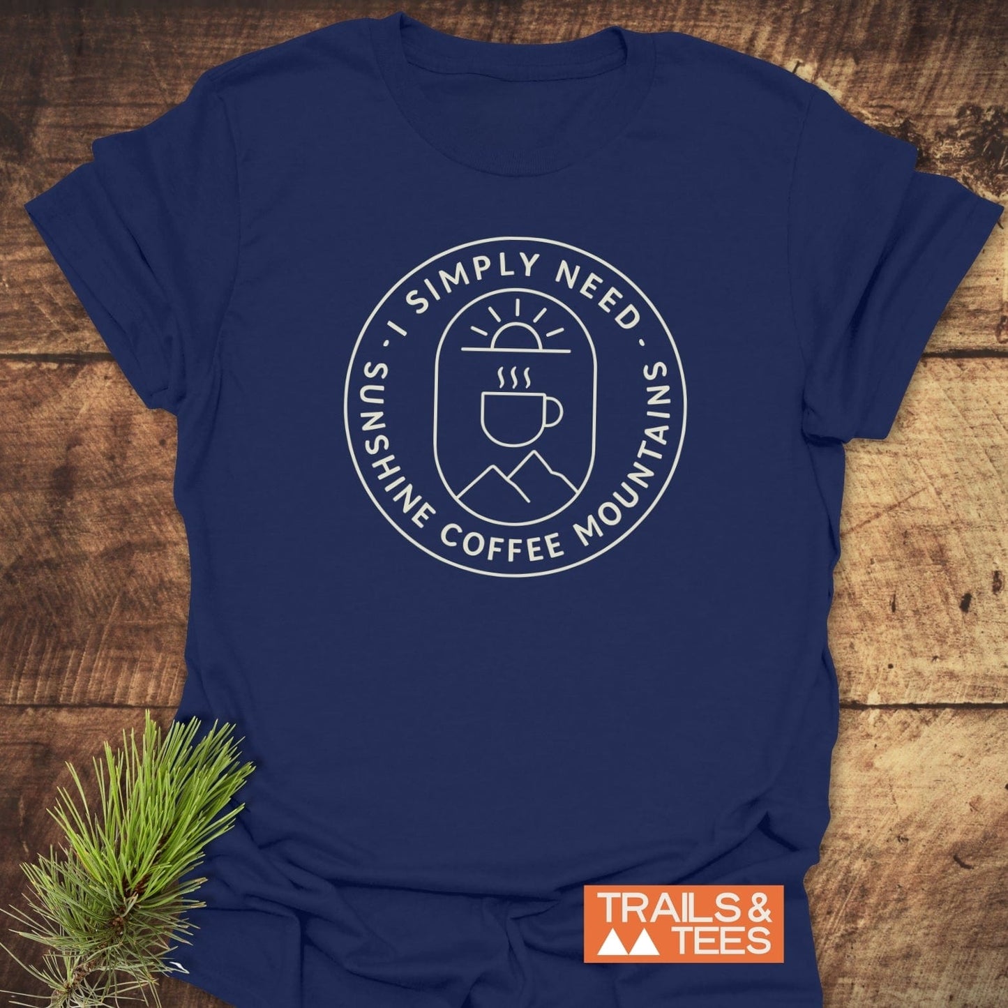 Minimalist Sunshine, Coffee & Mountains T-Shirt featuring a white circular graphic on navy, laid on wood with a pine sprig, brand label “TRAILS & TEES” visible.