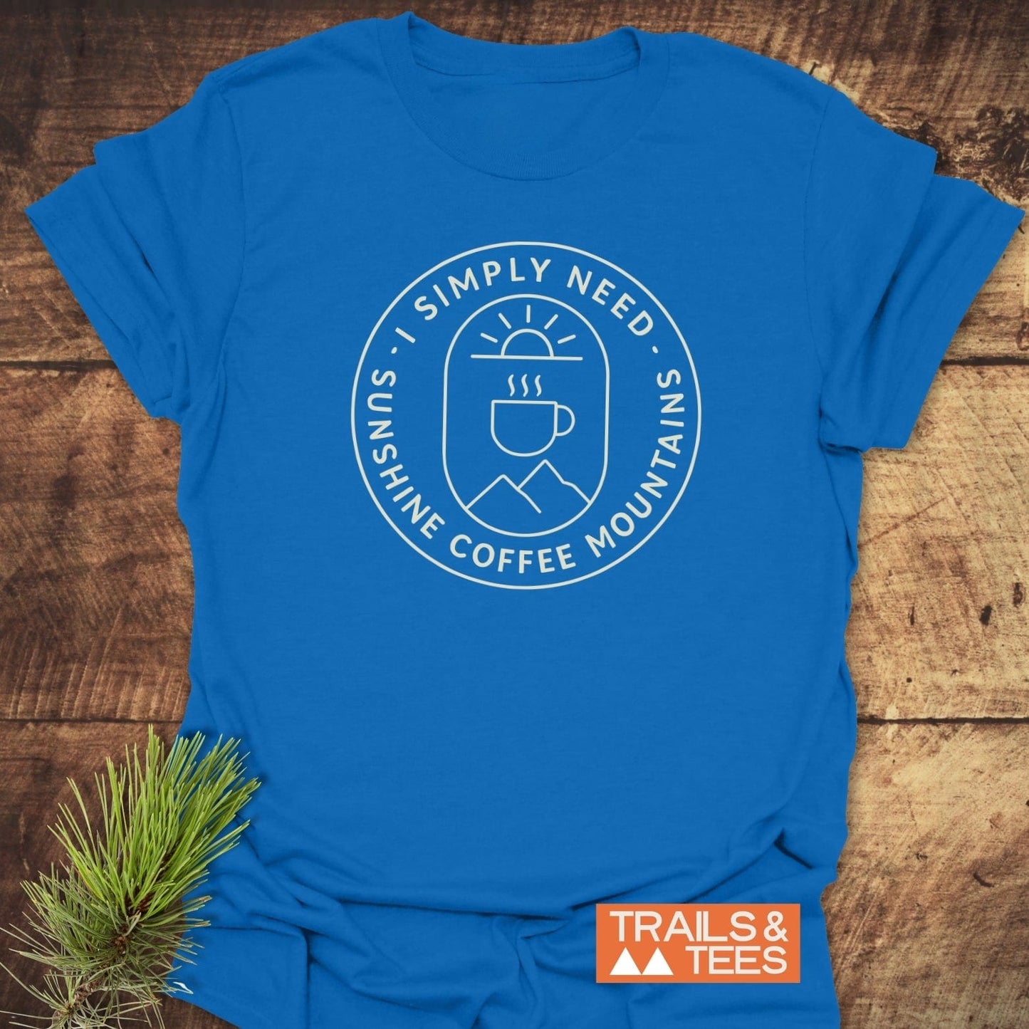 Minimalist Sunshine, Coffee & Mountains T-Shirt showcasing a white emblem with a sun, coffee cup, and mountains, laid on rustic wood, emphasizing outdoor adventure spirit.