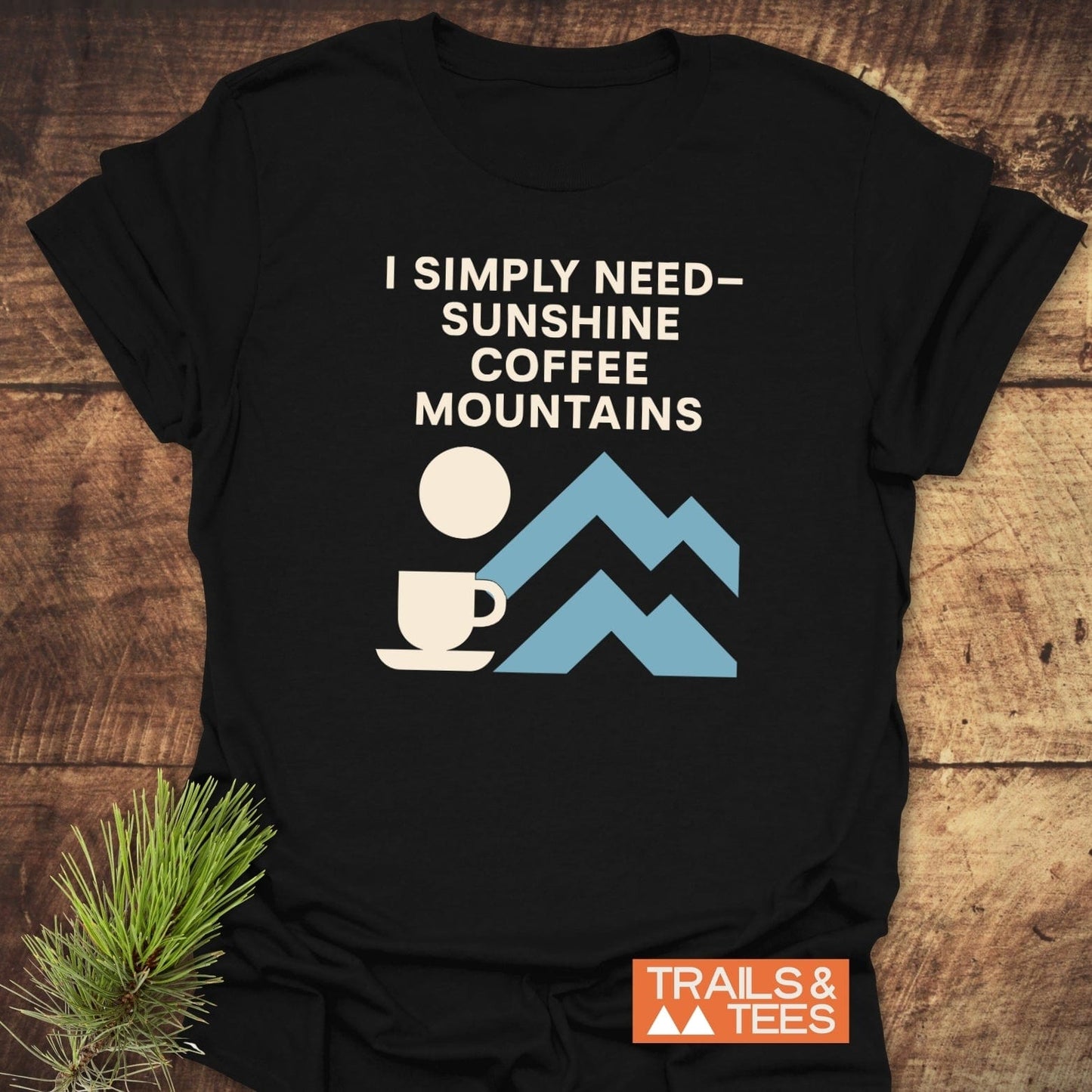 Geometric Mountain Love T-Shirt on wood surface, featuring a “I Simply Need” graphic with sun, coffee cup, and mountains. Includes Trails & Tees logo.