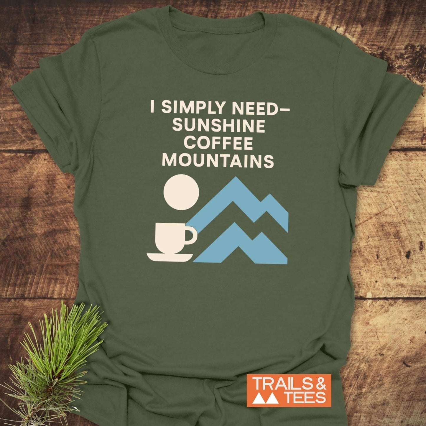 Geometric Mountain Love T-Shirt on wood, featuring “I SIMPLY NEED—SUNSHINE COFFEE MOUNTAINS” and minimalist sun, coffee, mountains graphic; Trails & Tees logo visible.