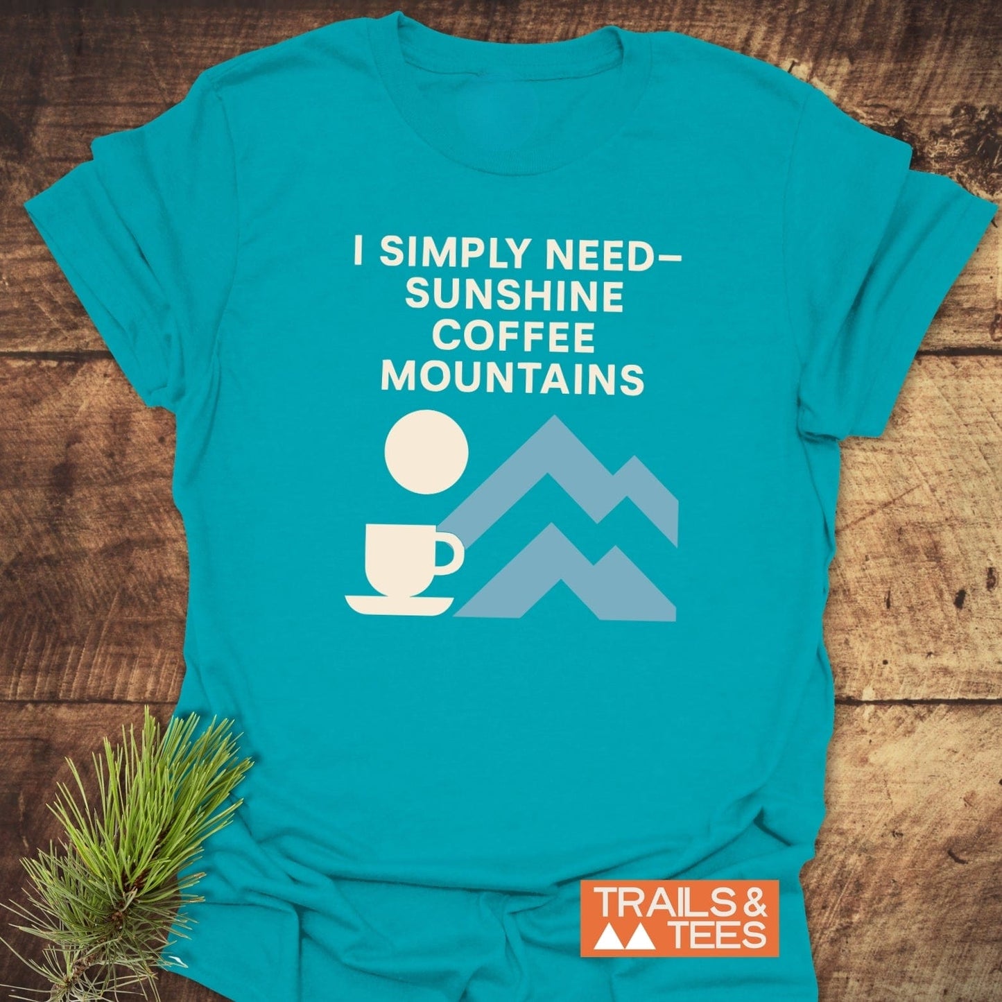 Geometric Mountain Love T-Shirt featuring an outdoorsy slogan with sun, coffee, and mountain icons. Laid on rustic wood with a pine sprig.