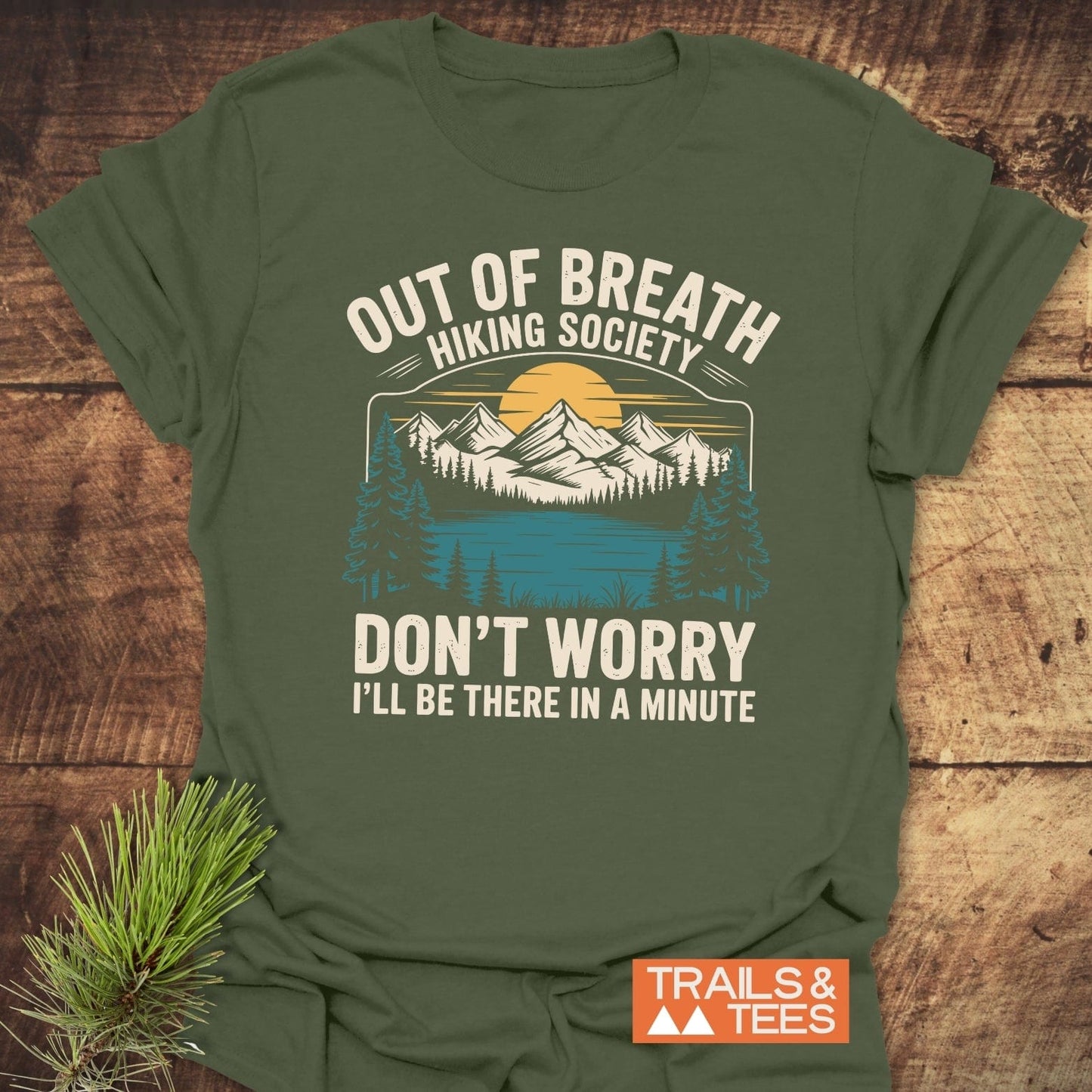 Forest-green Out Of Breath Funny T-Shirt with mountain graphic, styled on wood. Includes TRAILS & TEES logo, emphasizing adventure and durability.
