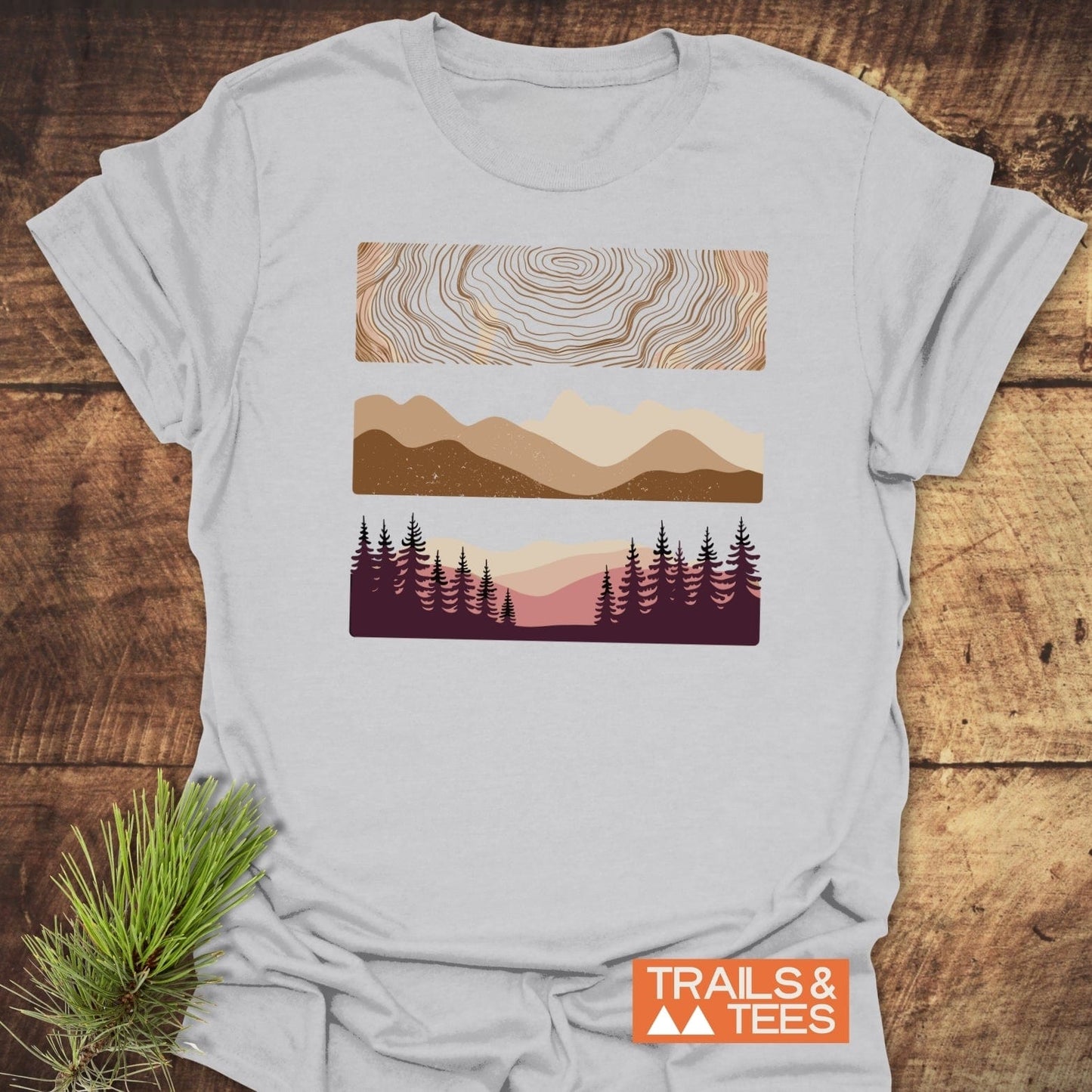Minimalist Mountains Graphic T-Shirt on wooden surface, featuring topographic lines, mountain silhouettes, and a forest scene. Includes TRAILS & TEES logo in corner.