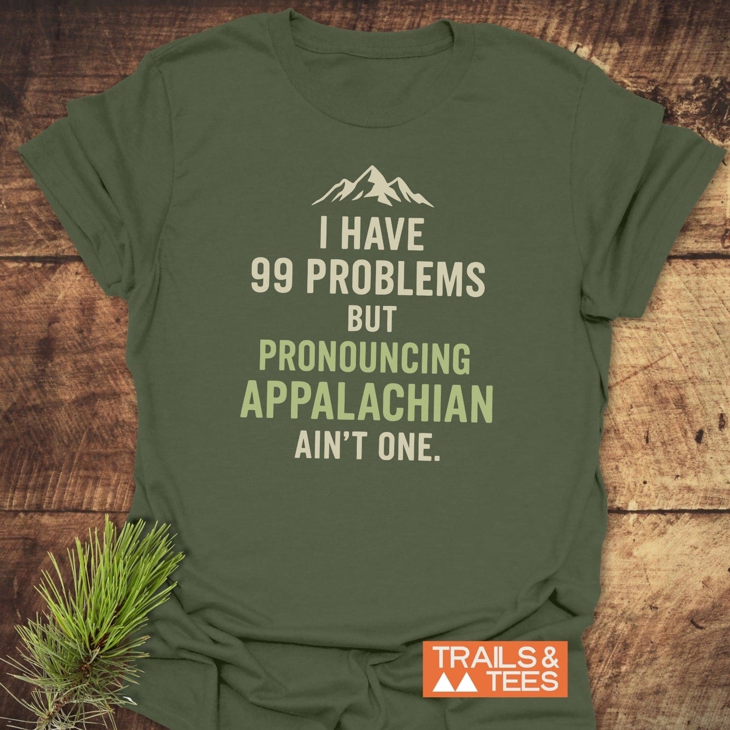 Olive-green 99 Problems Funny T-Shirt with Appalachian slogan, mountain graphic, and Trails & Tees logo, laid on wood with pine sprig accent.