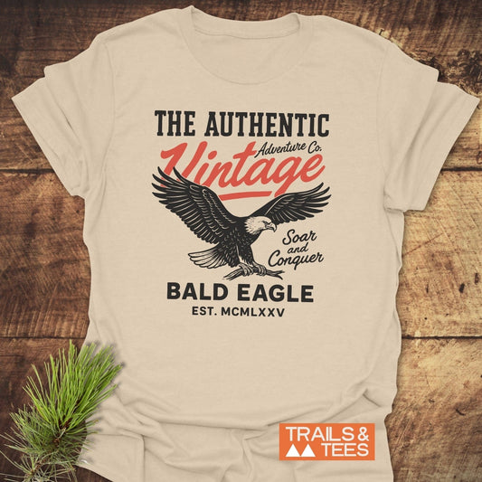 Vintage Bald Eagle T-Shirt featuring a bold eagle design and Soar and Conquer text, laid on wood with pine sprig accentuating its adventure theme.