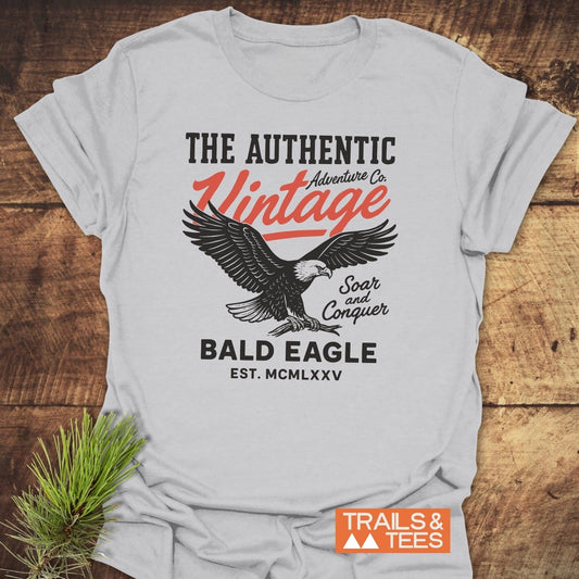 Vintage Bald Eagle T-Shirt featuring a bald eagle illustration and adventure-themed text, laid on wood with a pine sprig, showcasing outdoor-inspired design and comfort.