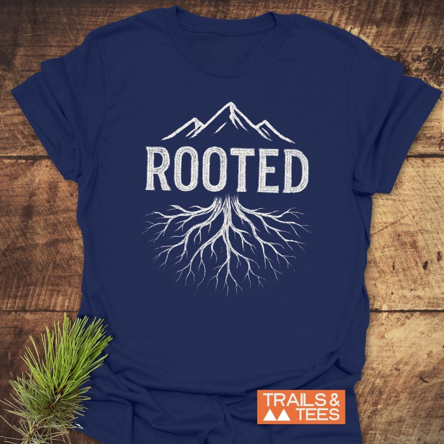 Rooted Mountain T-Shirt with a mountain and roots design, featuring the Trails & Tees logo, laid flat on a wooden surface.