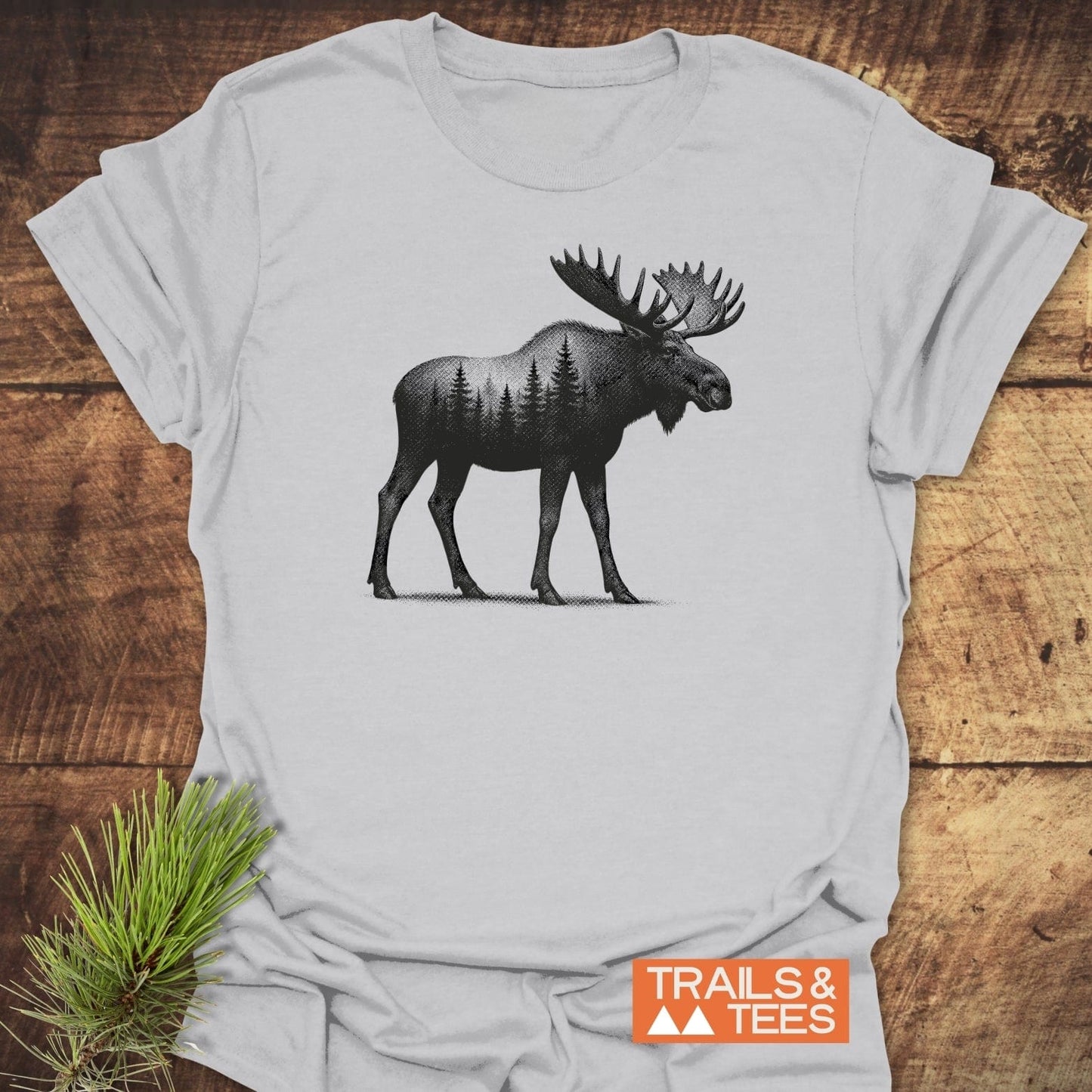 Moose Halftone T-Shirt featuring a moose silhouette with evergreen trees, laid flat with Trails & Tees logo and pine sprig, highlighting outdoor-inspired design.