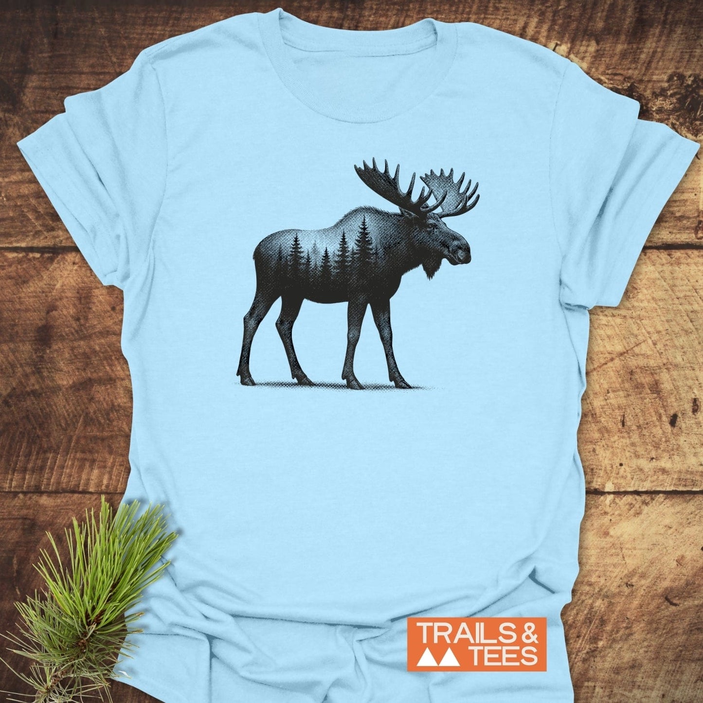 Moose Halftone T-Shirt featuring a black moose graphic with forest silhouette, laid on wood, pine sprig beside, and TRAILS & TEES logo visible.