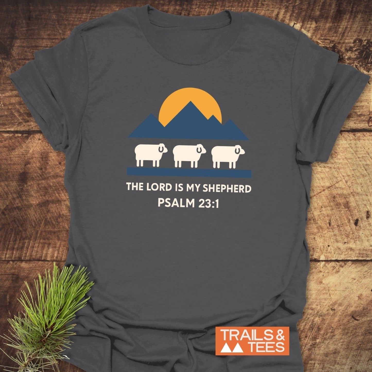 Charcoal-gray T-shirt with The Lord Is My Shepherd Psalm 23:1 design featuring a sun, mountains, and sheep. Trails & Tees logo at corner.