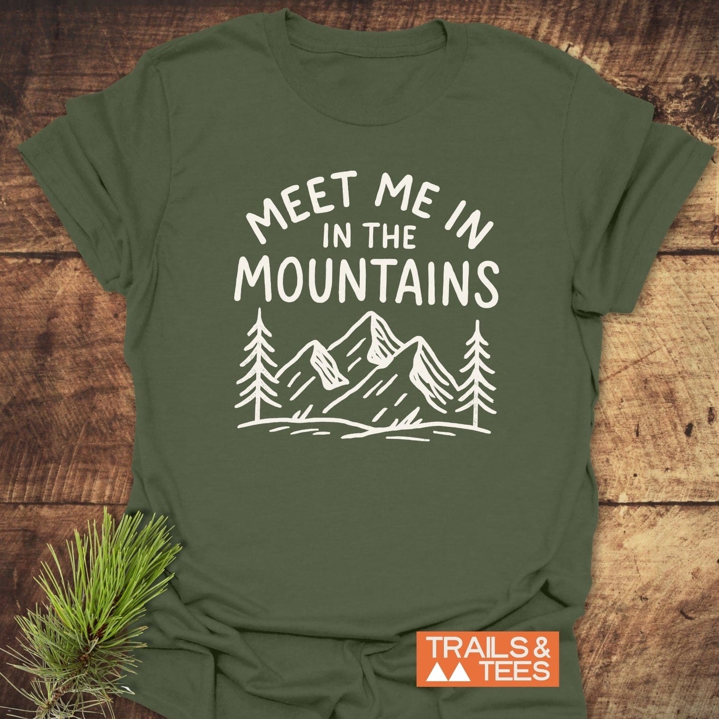 Meet Me In The Mountains T-Shirt featuring mountain peaks and pine trees graphic on soft, ring-spun cotton, laid on wood with pine sprig beneath.
