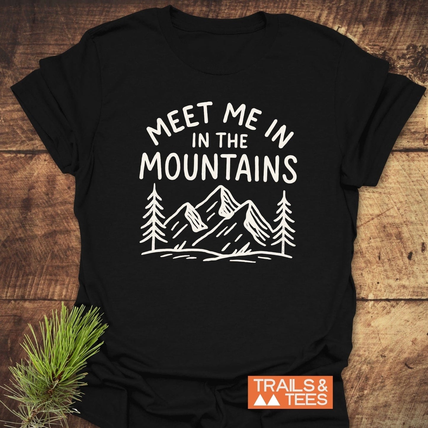 Meet Me In The Mountains T-Shirt with mountain and pine graphic, emphasizing outdoor adventures and comfort, displayed with natural elements for an outdoorsy feel.