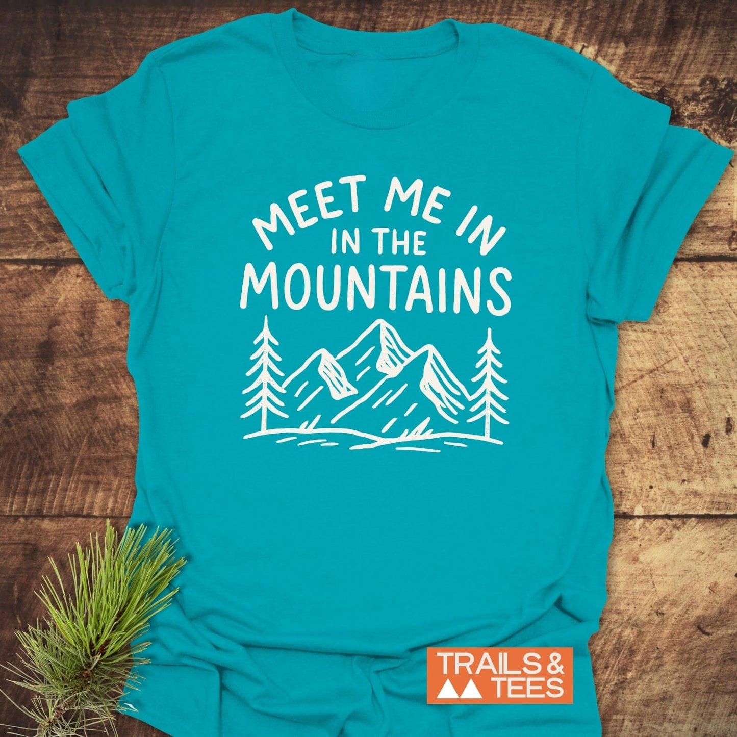Meet Me In The Mountains T-Shirt with white pine tree and mountain graphic, laid on wood. Features soft cotton, durable print, perfect for outdoor enthusiasts.