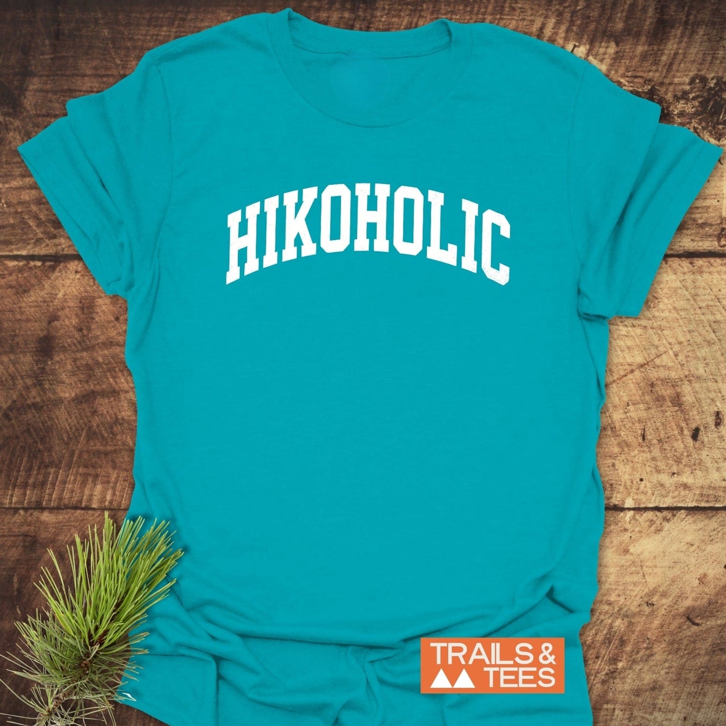 Hikoholic T-Shirt featuring white collegiate lettering on soft, lightweight cotton, laid on rustic wood with decorative evergreen and TRAILS & TEES logo.