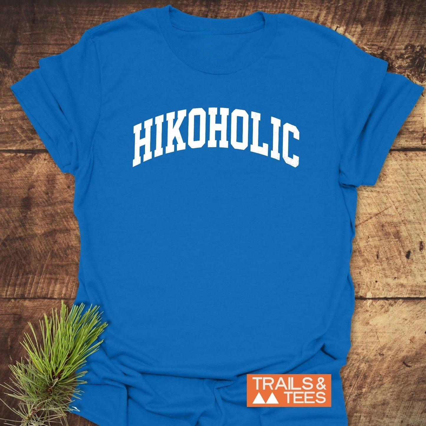 Hikoholic T-Shirt on rustic wood, featuring white collegiate-style text and orange TRAILS & TEES logo, accompanied by a pine sprig, highlighting outdoor adventure spirit.
